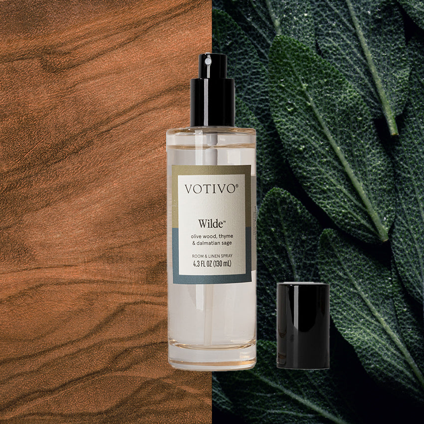 A clear bottle of Votivo Wilde Room & Linen Spray with a black cap is set against rich wood grain and green sage leaves, reflecting its signature rosemary sage fragrance.