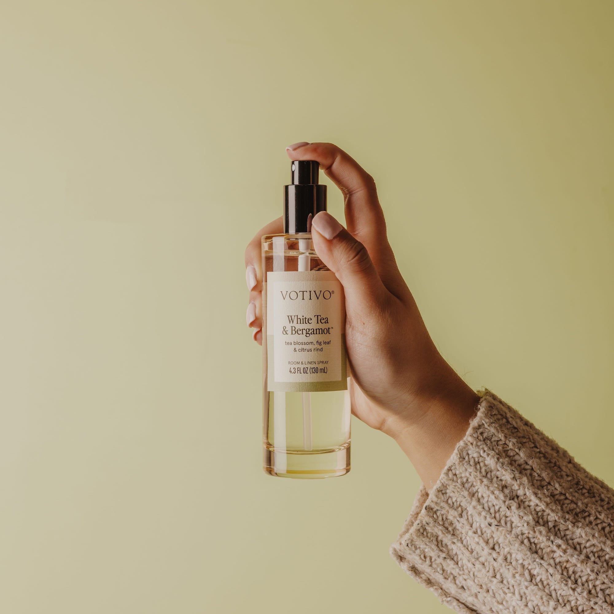 A hand in a beige knitted sleeve holds a clear Votivo White Tea & Bergamot Room & Linen Spray, fabric safe, against a light yellow-green background.