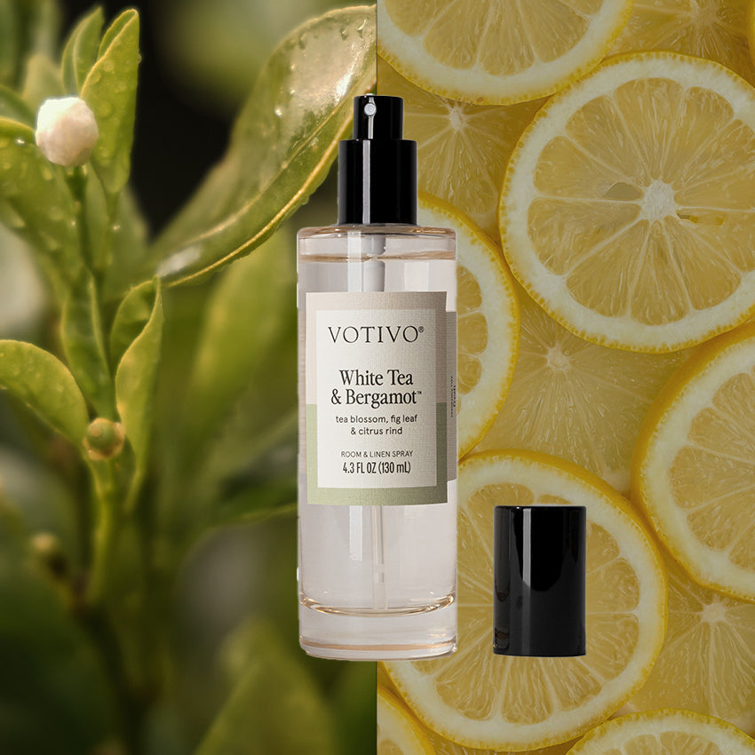 A clear Votivo White Tea & Bergamot Room & Linen Spray, cap off, rests against green tea leaves and sliced lemons—perfect for safely refreshing fabric with its fragrance.
