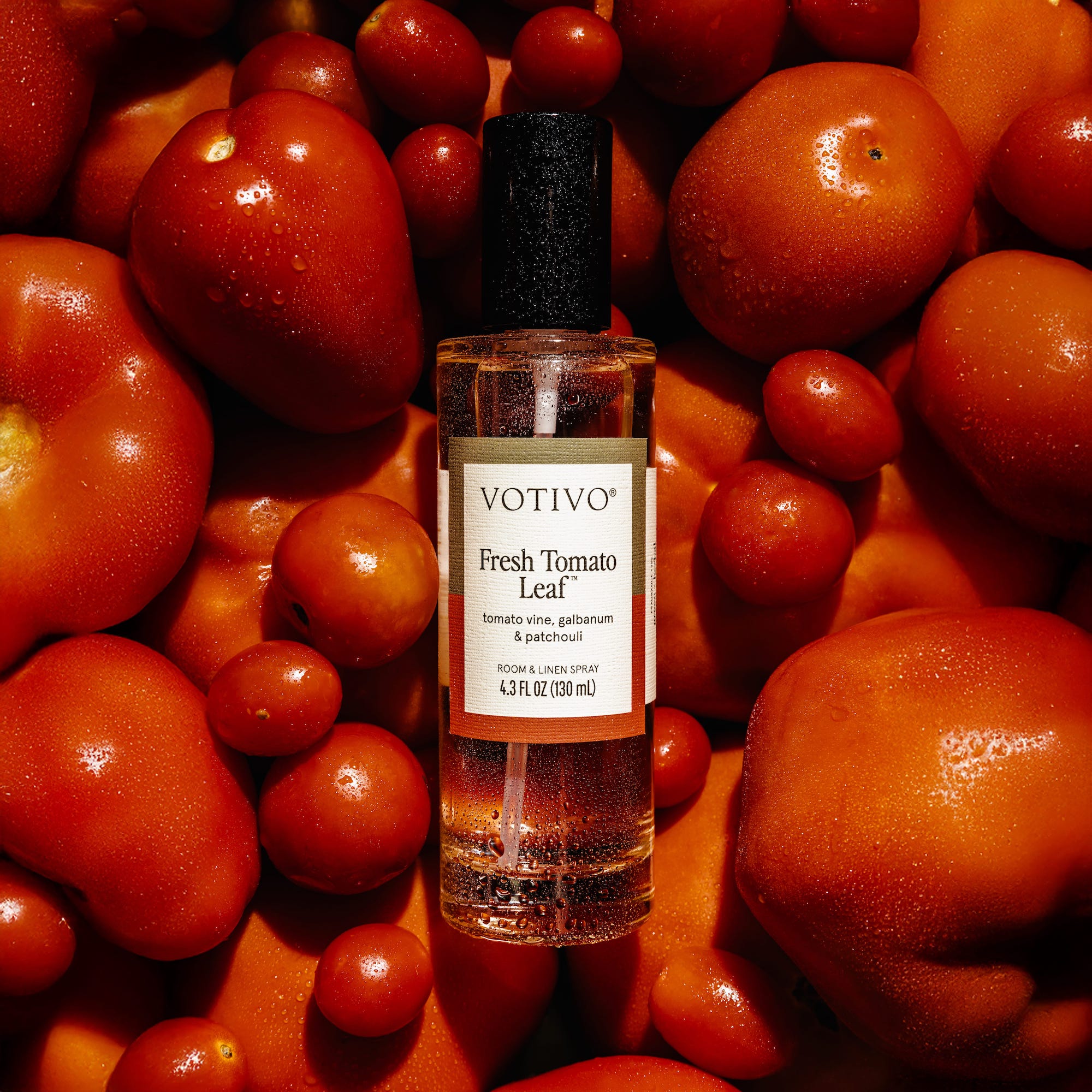 A clear bottle of Votivo Fresh Tomato Leaf Room & Linen Spray with a black cap and white label sits among dewy, ripe red tomatoes, highlighting its fabric-safe and refreshing qualities.