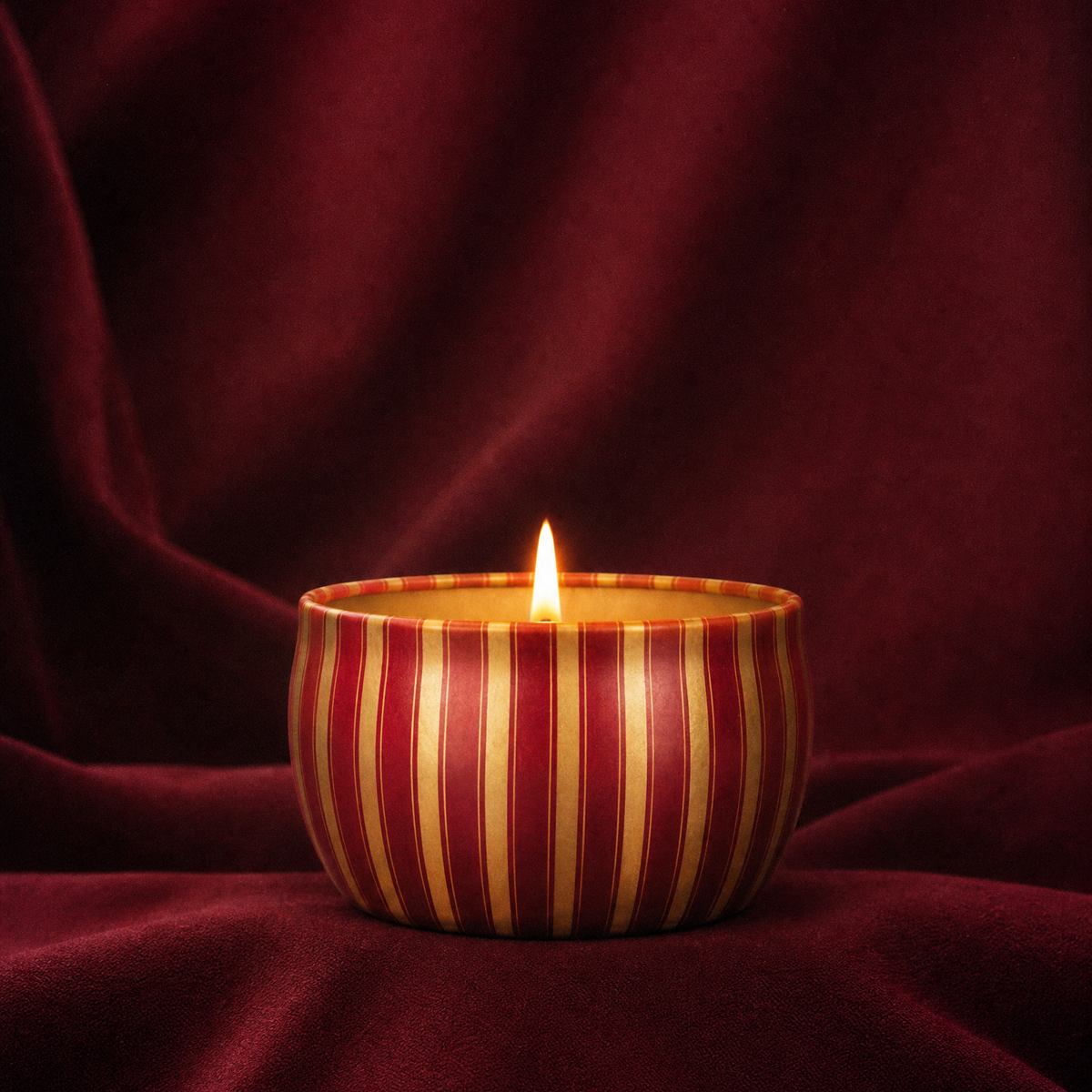 A lit Velvet Rouge tin candle with striped packaging against a velvet burgundy backdrop