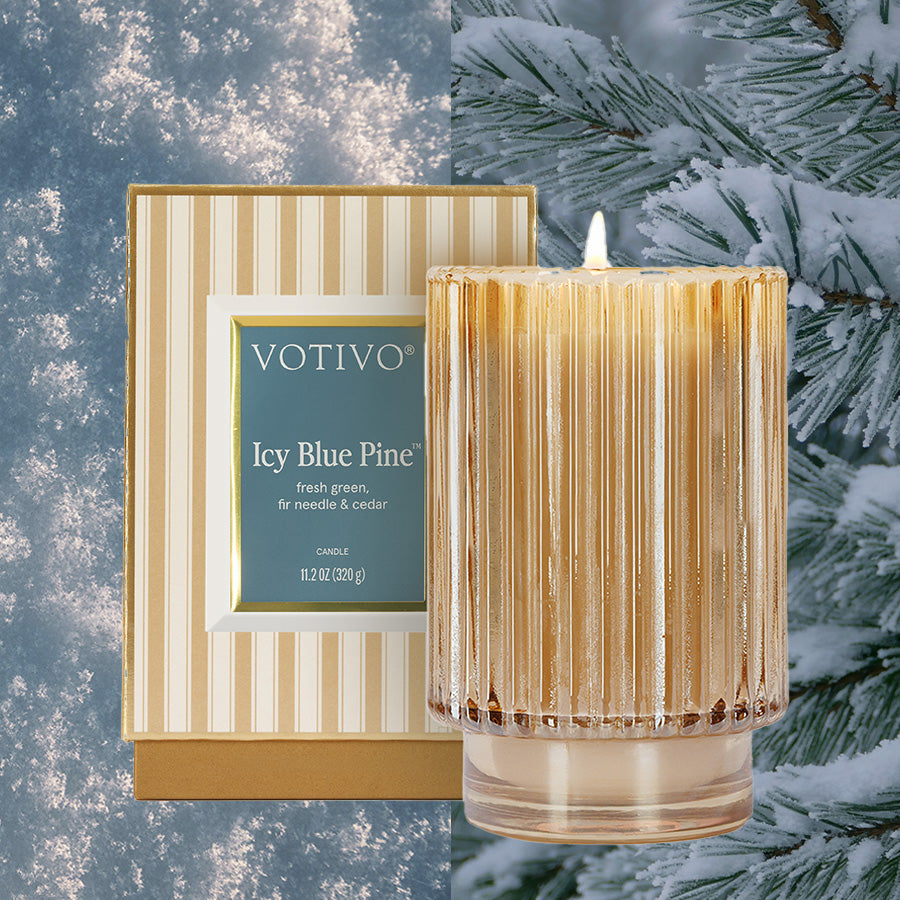 The Votivo Icy Blue Pine Holiday Candle, crafted from a soy blend wax, rests in a ribbed glass container next to its striped box, accented by snow-dusted pine branches in the background.