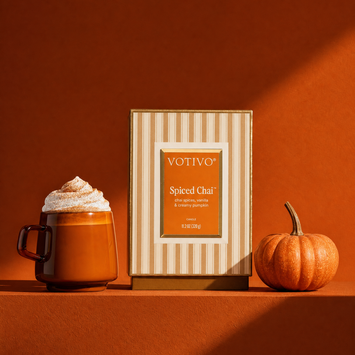 Votivo Spiced Chai candle
box standing on a warm terracotta surface, flanked by a whipped-cream-topped mug and a small pumpkin, in a cohesive autumn color palette.