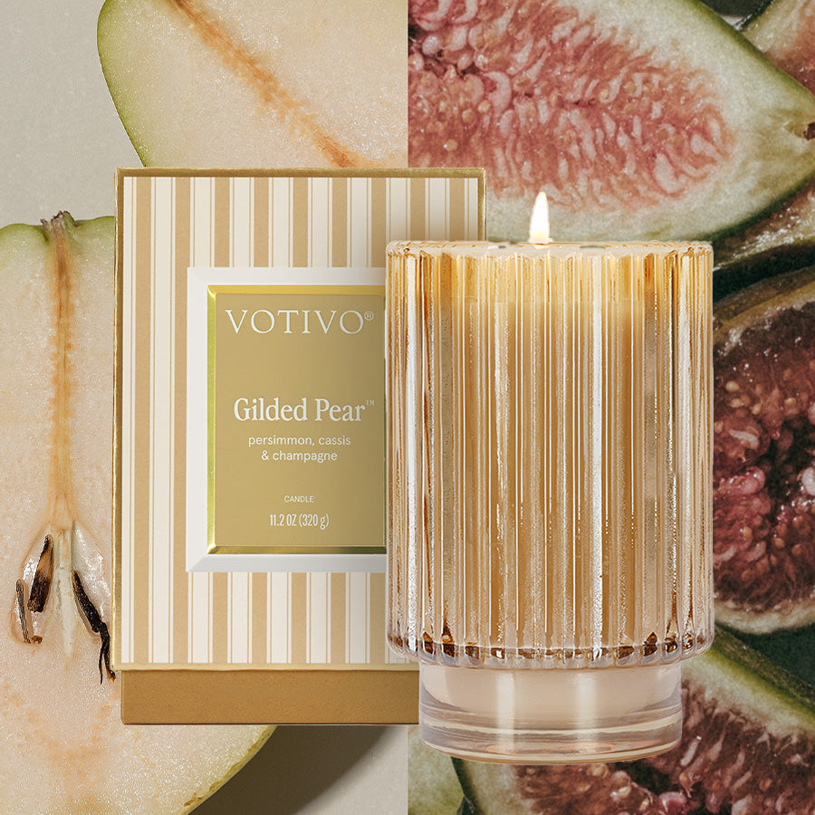 The Votivo Gilded Pear Holiday Candle, crafted from a soy blend and presented in a ribbed glass vessel, is lit beside its gold and cream striped box, with sliced pears and figs adding festive flair in the background.