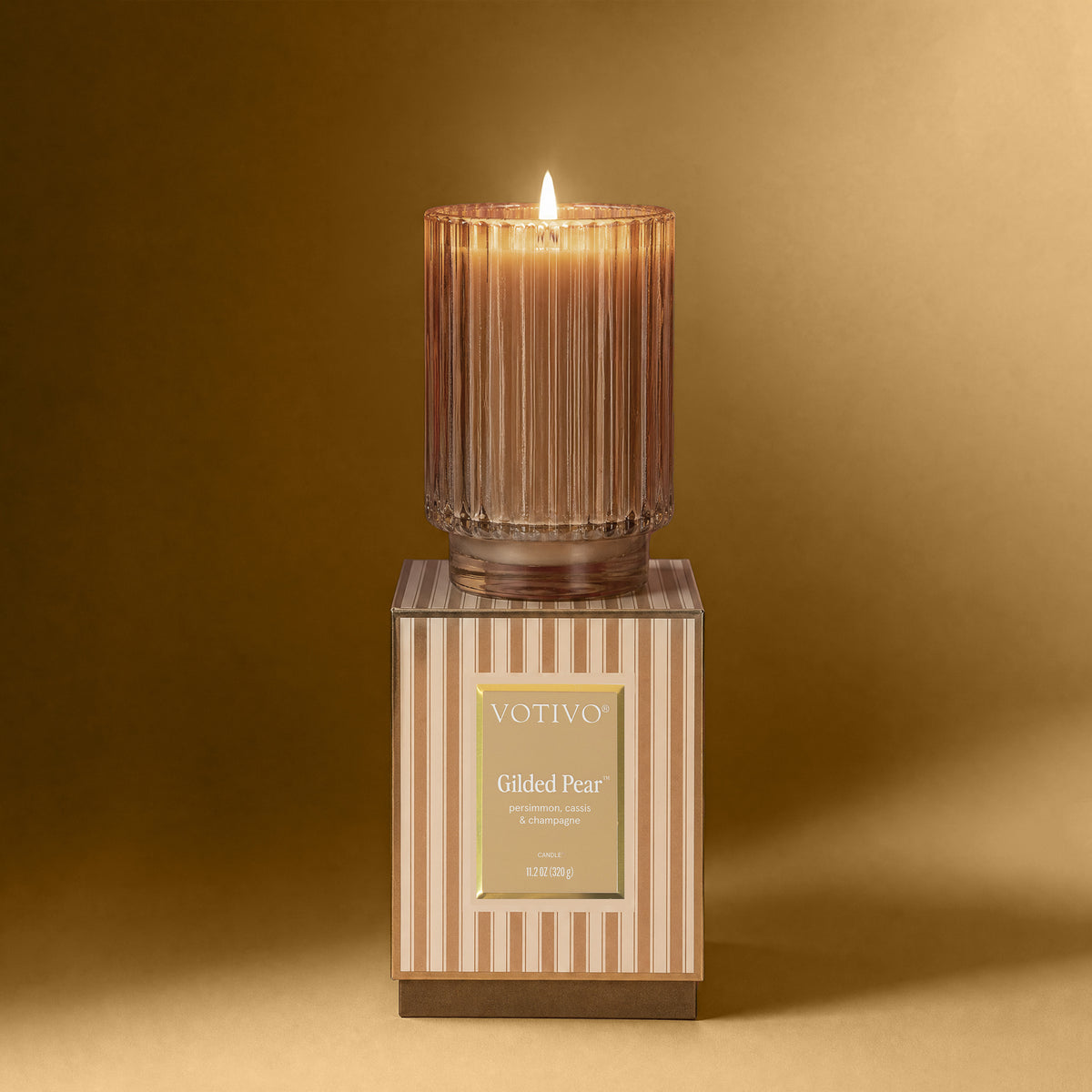 A lit Gilded Pear ribbed candle vessel sitting on a striped box against a gold background
