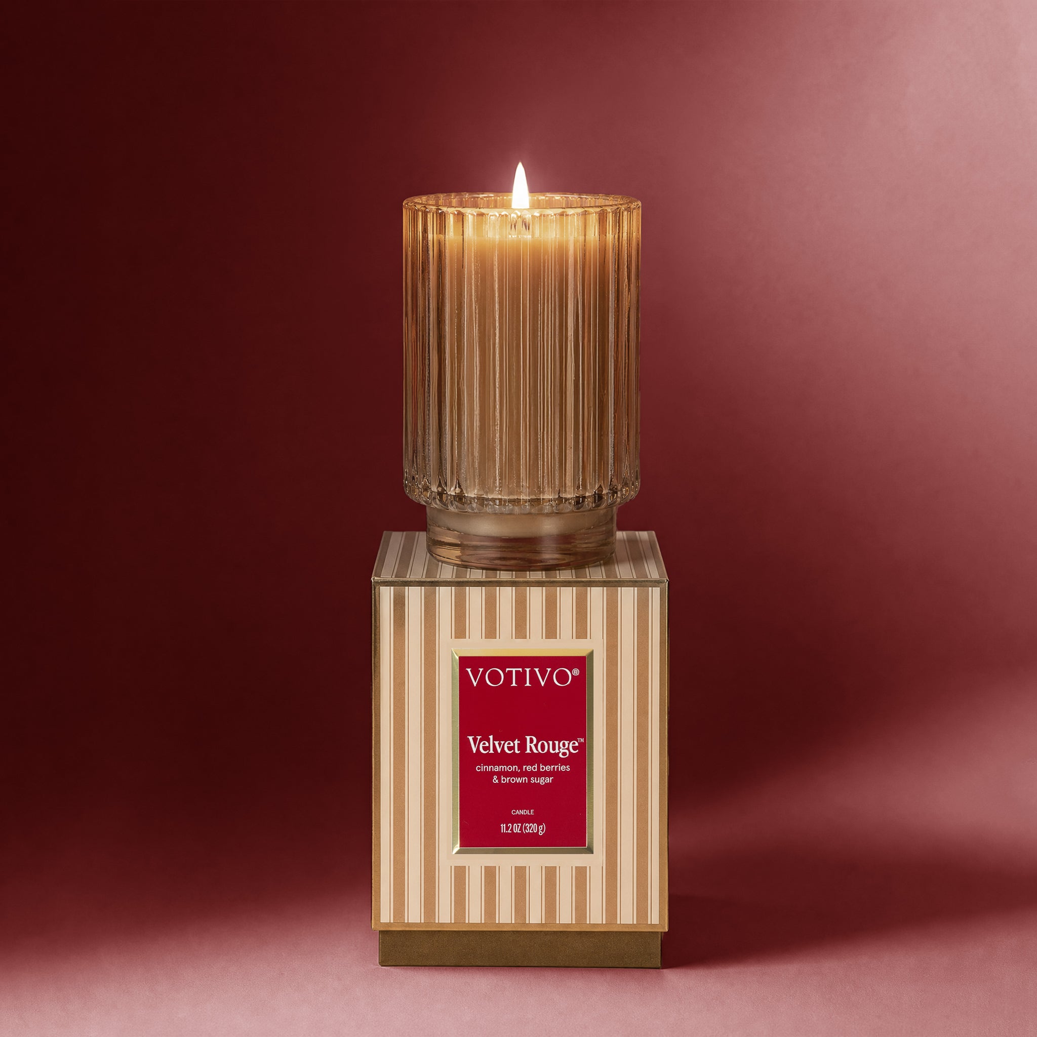 A lit Votivo Velvet Rouge Holiday Candle rests atop its coordinating striped box against a red backdrop. Housed in a ribbed glass jar and made from soy blend wax, the candle's label features fragrance notes of cardamom, red berries, and creamy woods.