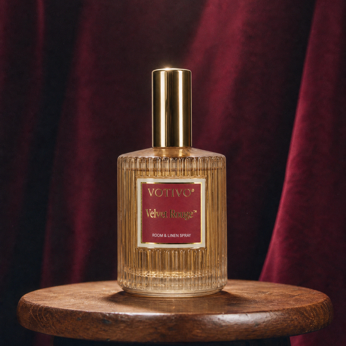 A Votivo Velvet Rouge Room & Linen Spray sitting on a wood stool against a burgundy background