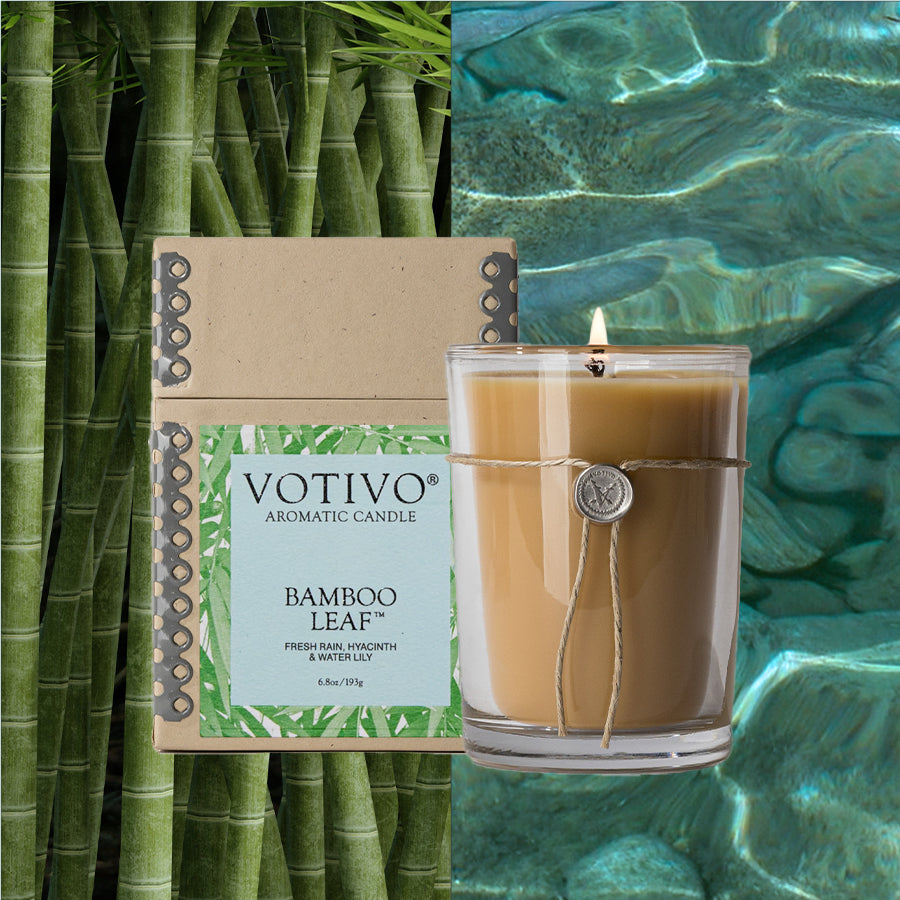 The Votivo Bamboo Leaf 6.8 oz. Aromatic Candle, in a twine-wrapped glass and soy blend wax, is shown with its box against a backdrop of bamboo stalks on the left and clear blue water on the right.