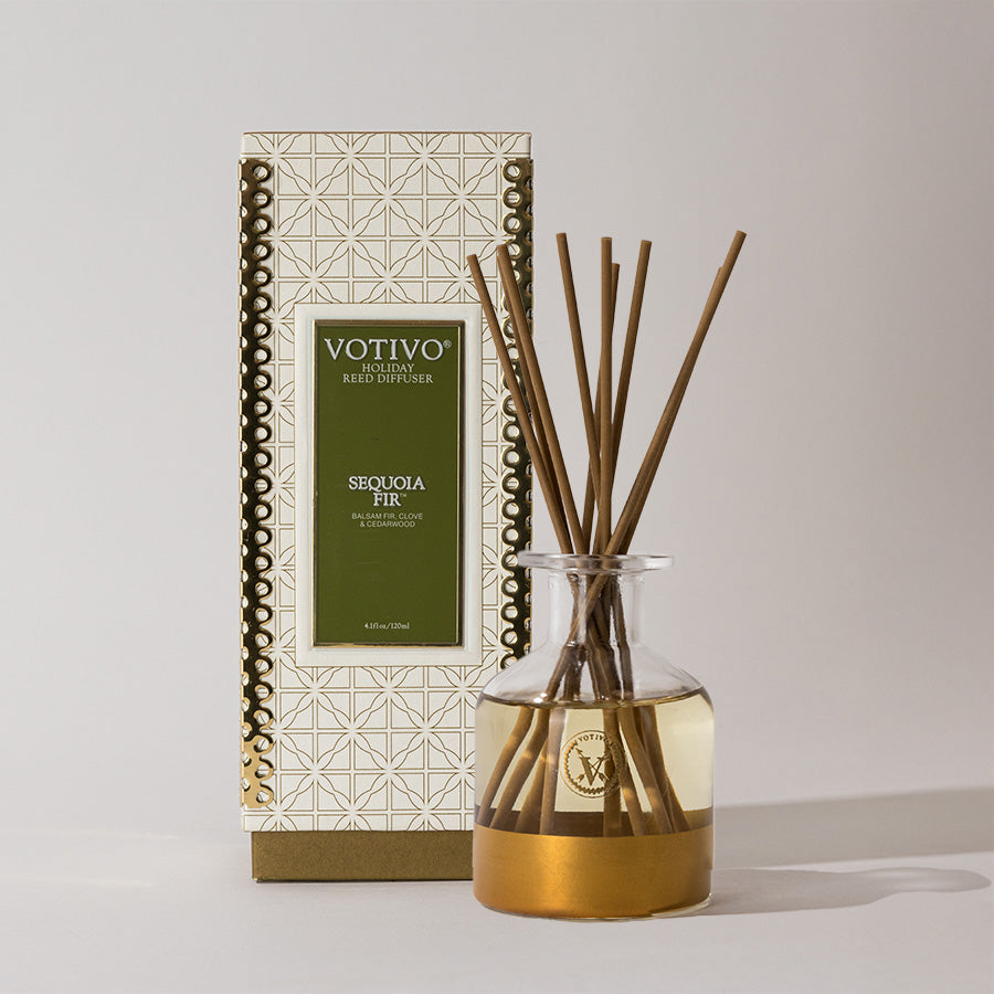 The Votivo Sequoia Fir Holiday Reed Diffuser includes a clear glass bottle with golden fragrance and brown rattan reeds, displayed in front of its decorative box featuring a geometric pattern and green label.