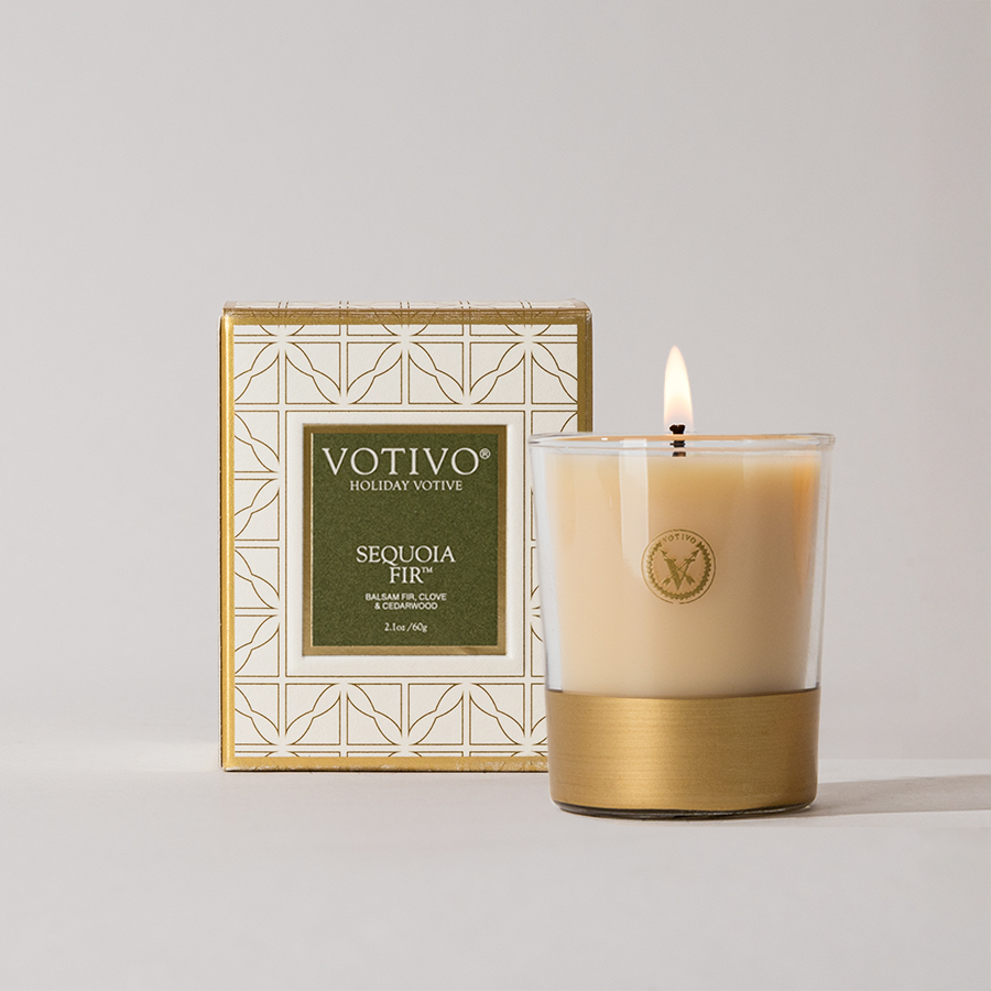 The Votivo Sequoia Fir Holiday Votive candle glows in a glass holder with a gold base, placed before its decorative box featuring a geometric gold and white design.