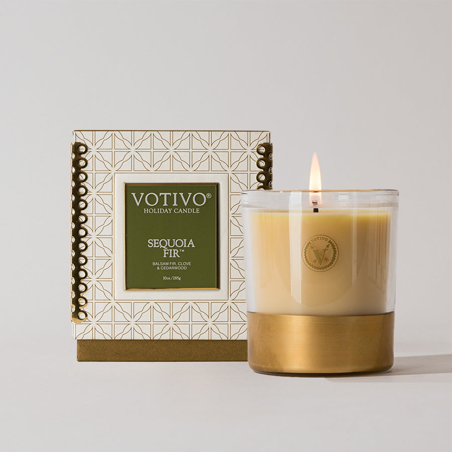 The Votivo Sequoia Fir 10 oz. Holiday Candle, made with a soy blend and set in a glass container with a gold base, is lit beside its geometric-patterned box, highlighting its festive scent against a plain white background.