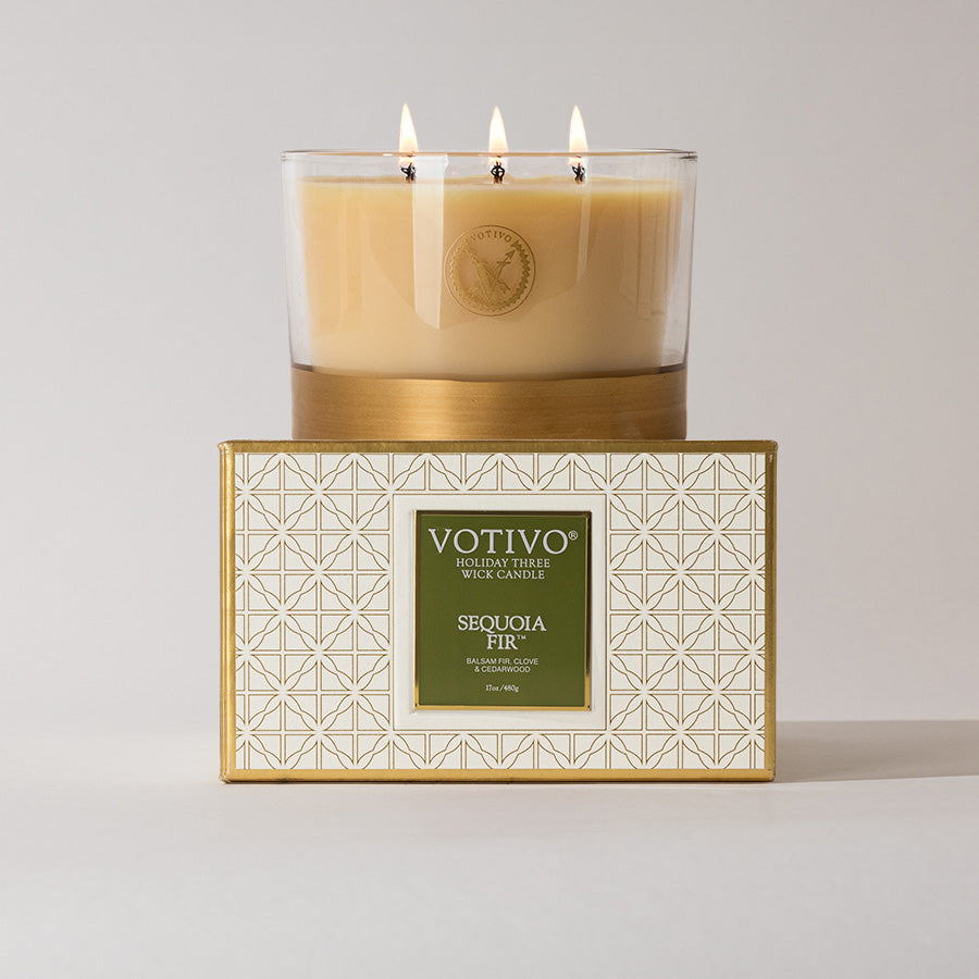 The Votivo Sequoia Fir Holiday 3 Wick Candle features a soy blend in a beige glass container, presented atop a white box with an elegant gold geometric pattern and labeled for the holiday season.