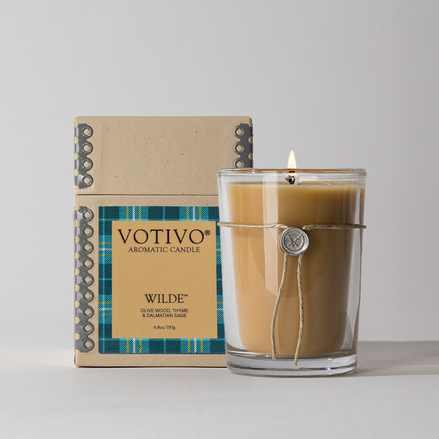 A lit Votivo Wilde 6.8 oz. aromatic candle in a clear glass holder, tied with string and a wax seal, sits next to its beige and blue plaid box labeled with olive wood, thyme, and balsam & sage notes.