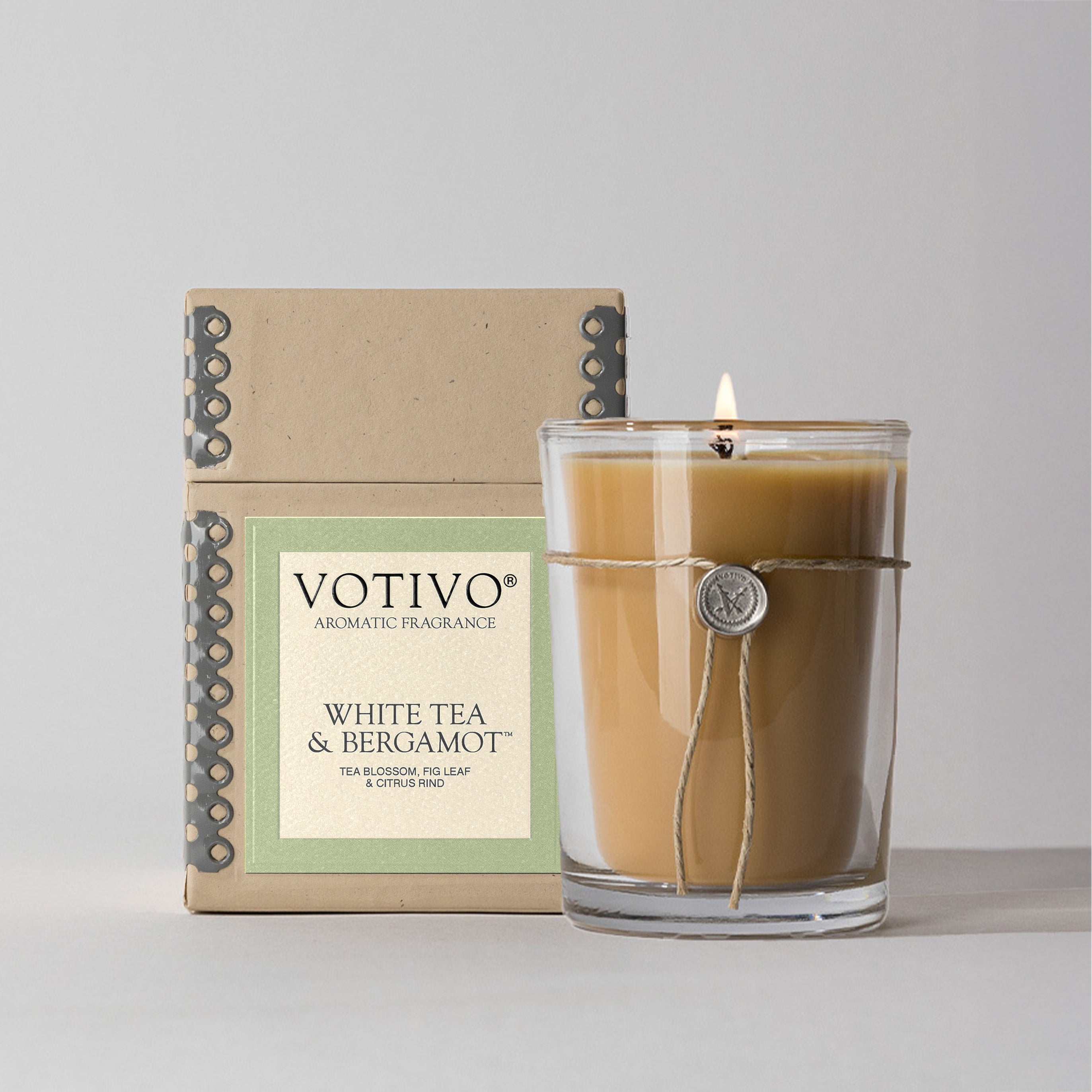 The Votivo White Tea & Bergamot 6.8 oz aromatic candle, made with soy blend wax and finished with a wax seal charm, glows next to its kraft paper box packaging with matching labels and decorative stitching.