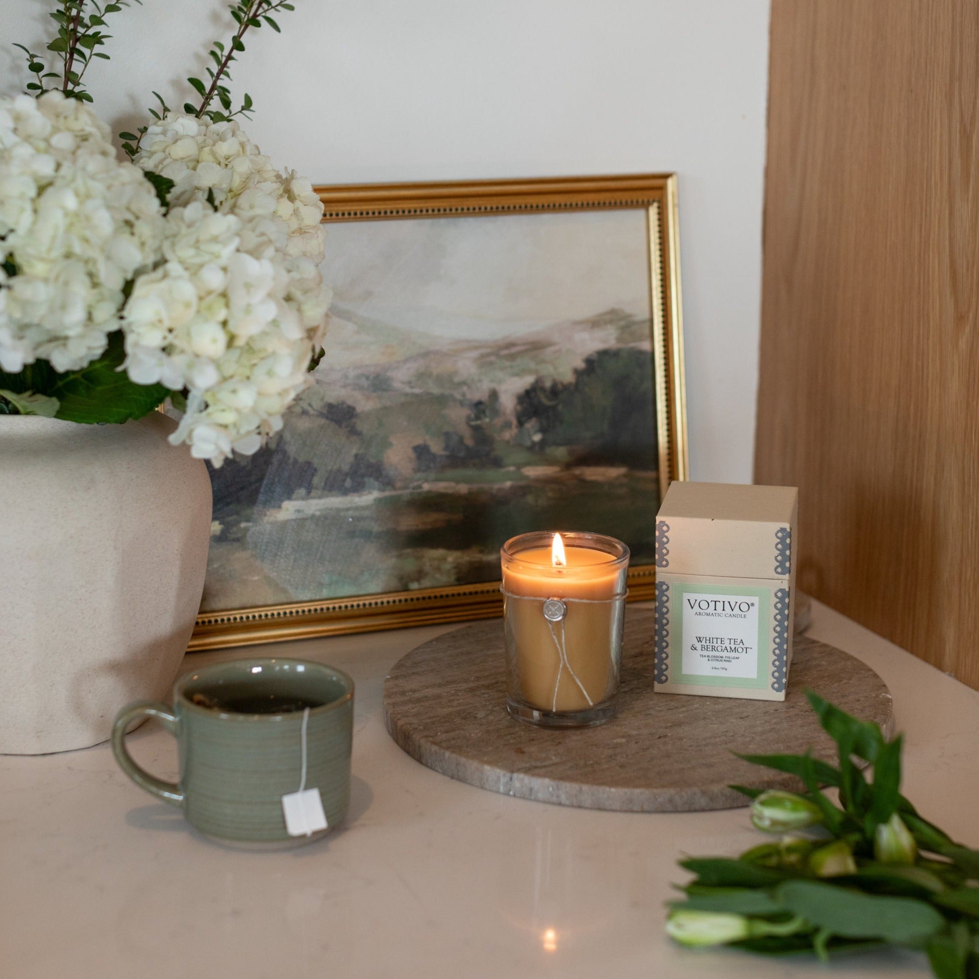 A Votivo White Tea & Bergamot 6.8 oz Aromatic Candle, a green mug with tea, white flowers in a vase, and its box are arranged on a table near tulips and a landscape painting.