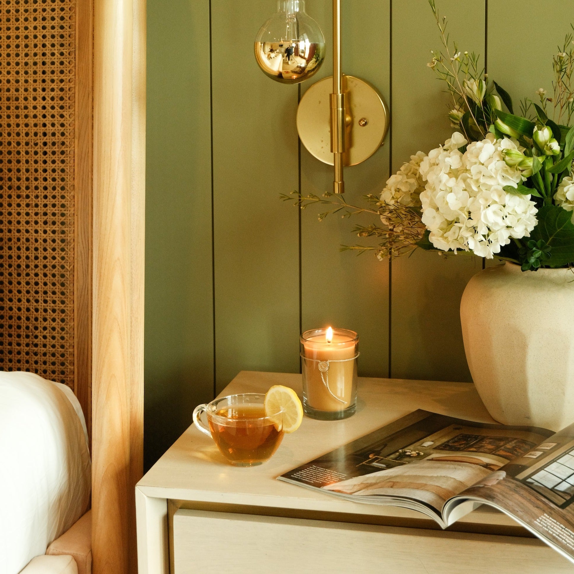 A cozy bedside table with a cup of tea garnished with lemon, a lit Votivo White Tea & Bergamot 6.8 oz Aromatic Candle, a large vase of white flowers, and an open magazine beside a bed with wood and rattan headboard and olive green wall.