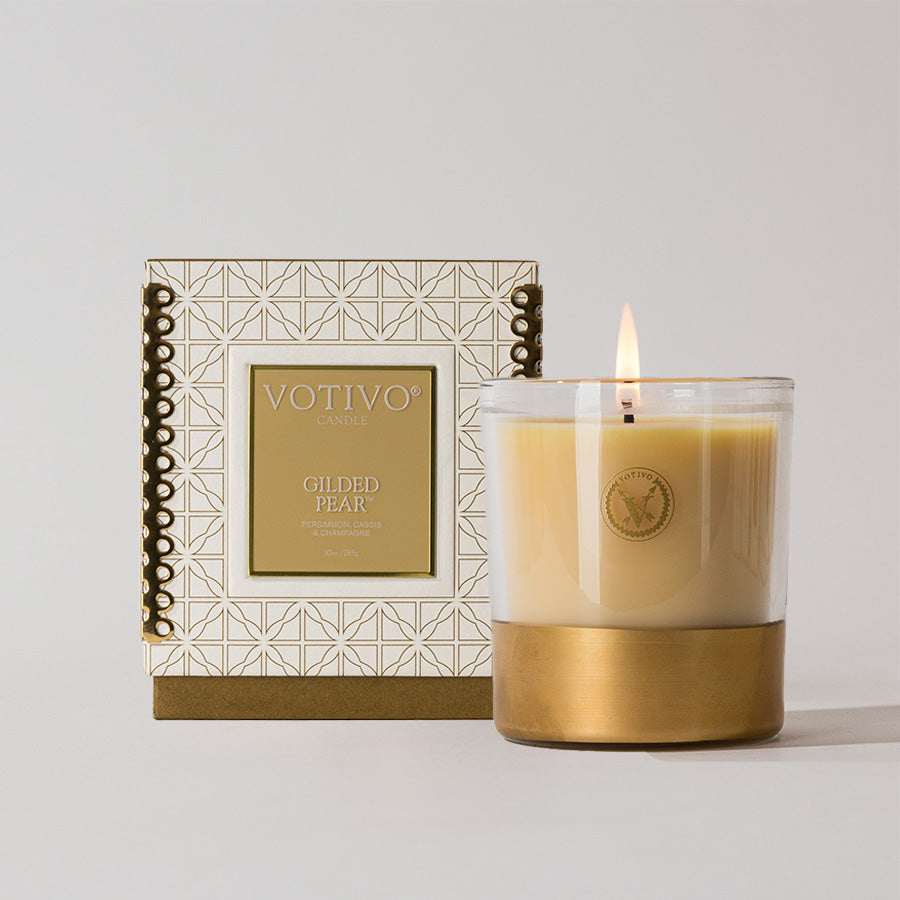 The Votivo Gilded Pear 10 oz. Holiday Candle, made with a soy blend and set in a clear glass jar with a gold base, sits in front of its decorative box adorned with geometric patterns and gold accents on a light background.