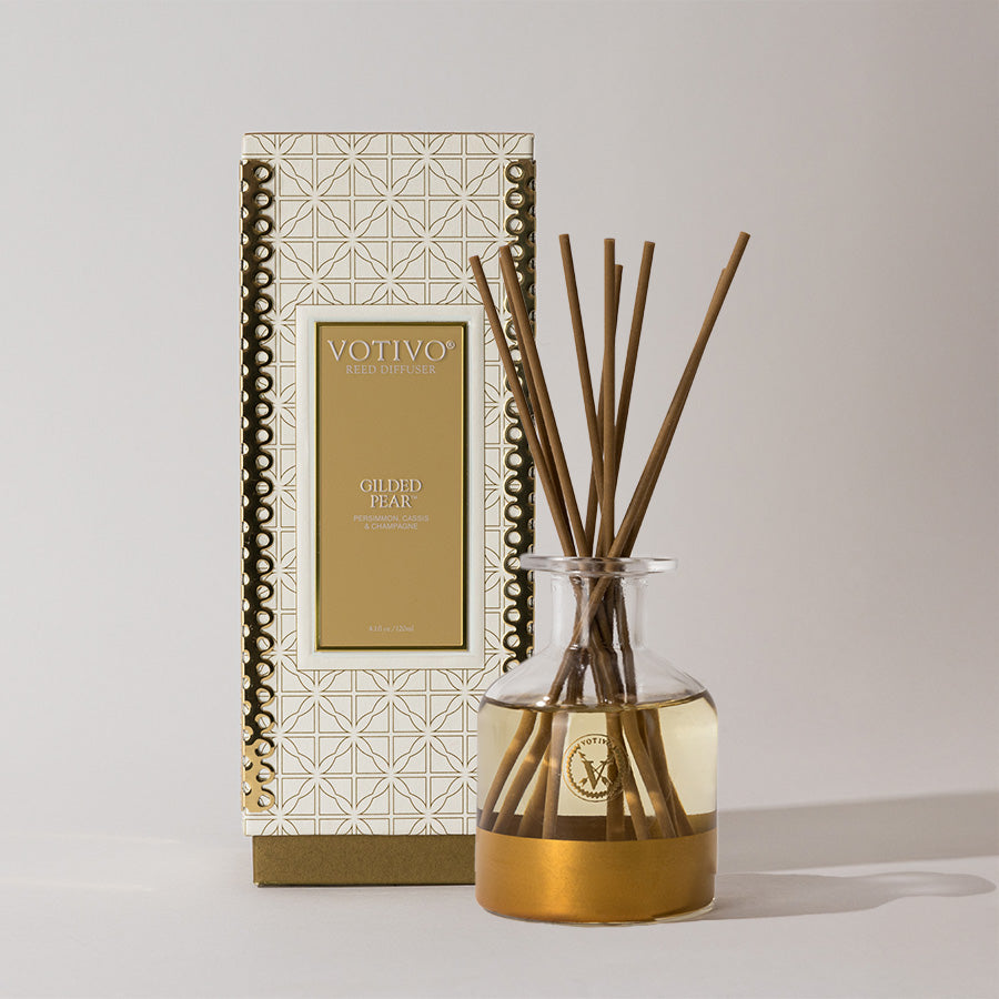 A Votivo Gilded Pear Holiday Reed Diffuser, in a gold-patterned box, features a glass bottle filled with fruity fragrance and thin reeds arranged inside.
