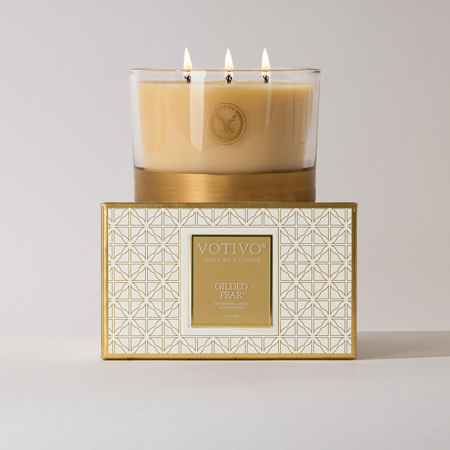 The Votivo Gilded Pear Holiday 3 Wick Candle sits atop its decorative white and gold box. Lit, it fills the plain background with a warm glow and the signature gilded pear fragrance.