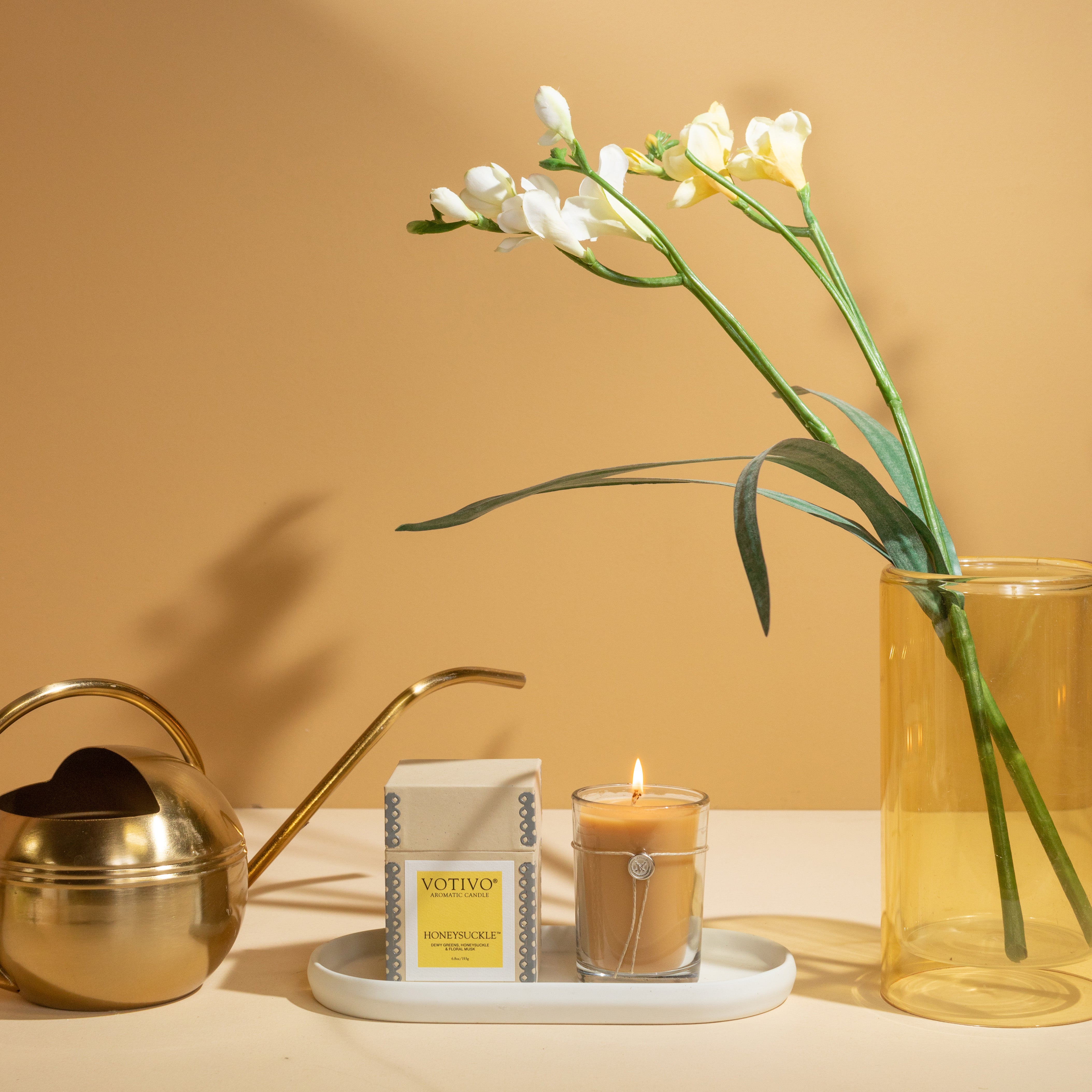 A gold watering can, a lit Votivo Honeysuckle 6.8 oz. Aromatic Candle, and a box labeled Votivo Honeycomb rest on a white tray beside a tall yellow vase with white flowers, all displayed against a beige background.