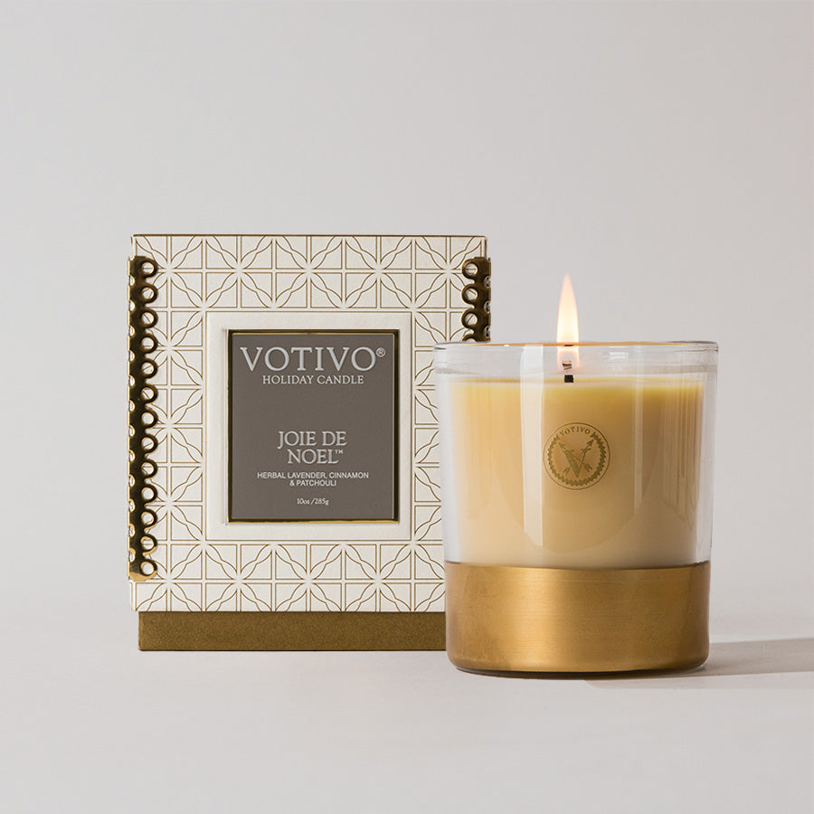 A lit Votivo Joie de Noel 10 oz. Holiday Candle in a glass jar with a gold base sits before its decorative white and gold box, featuring geometric patterns and a label with herbal lavender, cinnamon, and bergamot notes.