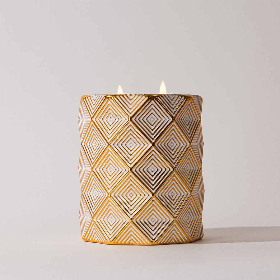 The Votivo Joie de Noel Holiday Decorative Candle, featuring two flames and soy blend wax, comes in a white and gold geometric diamond-patterned holder, perfect for adding festive elegance to any space.