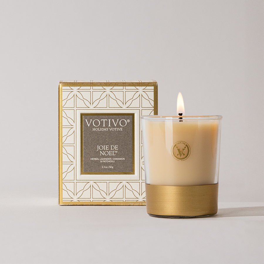 The Votivo Joie de Noel Holiday Votive, a luxury soy blend candle in a glass holder with gold base, sits before its white and gold box, filling the air with herbal, lavender, and cinnamon notes.