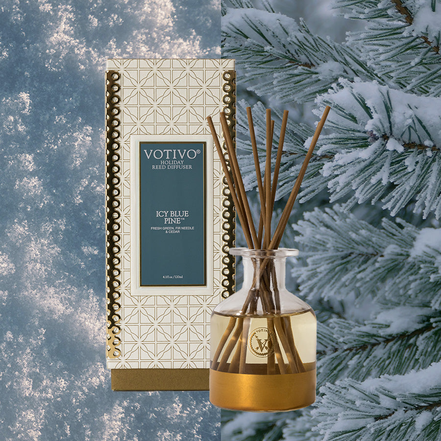 The Votivo Icy Blue Pine Holiday Reed Diffuser, in a gold and white geometric box, is shown alongside snowy pine branches. The glass bottle contains thin rattan reeds for festive fragrance.
