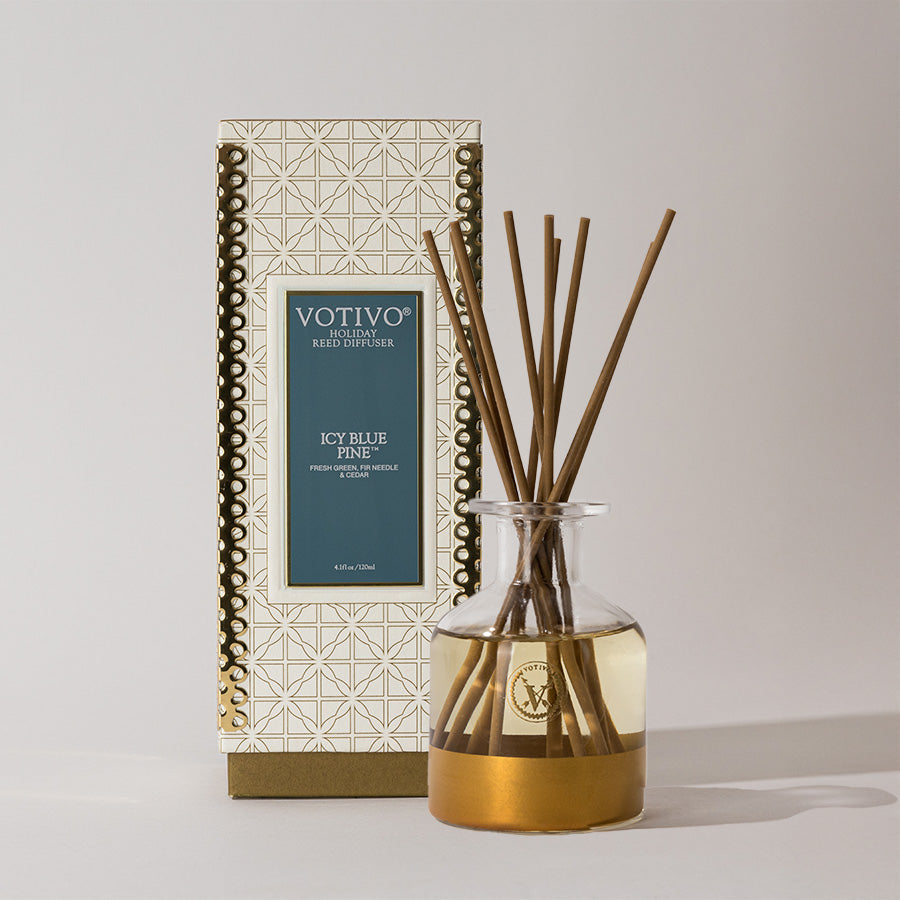 The Votivo Icy Blue Pine Holiday Reed Diffuser features brown reeds in a glass bottle of pale golden liquid, set before its geometric gold-and-white box, bringing a fresh scent to your space.