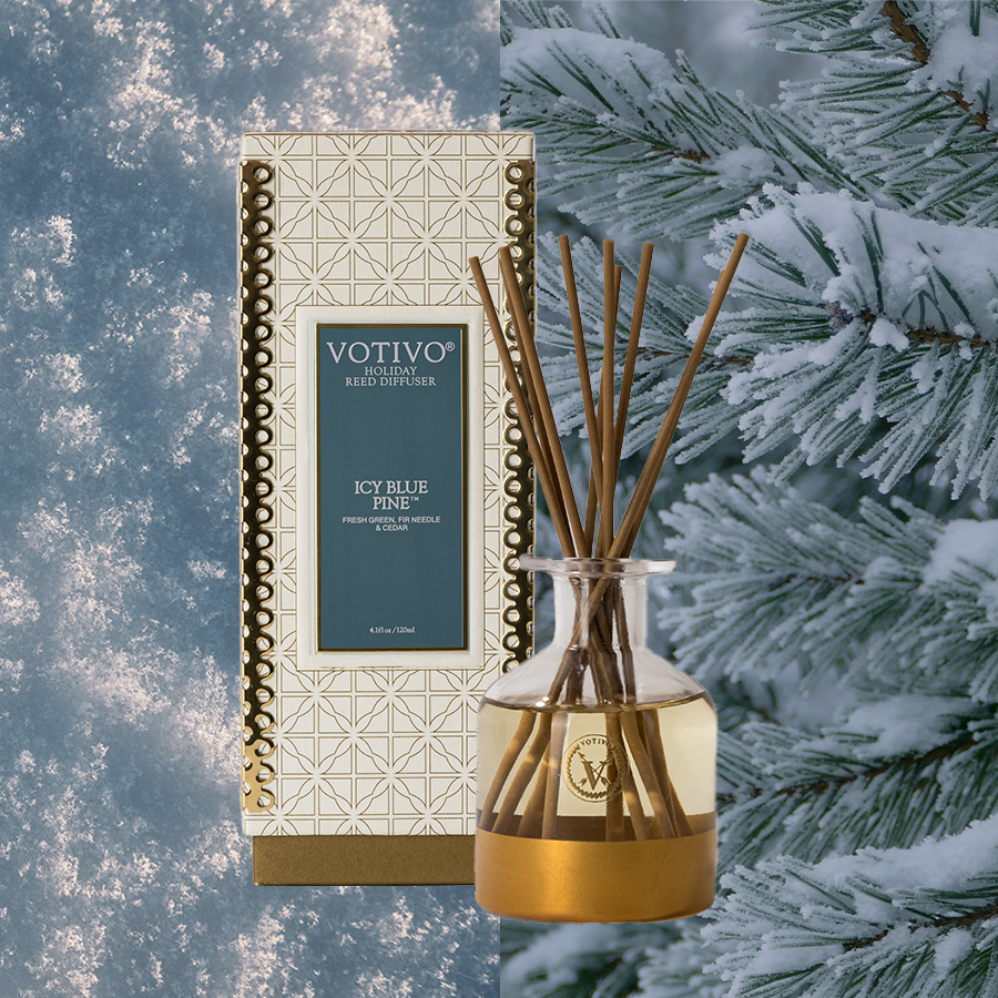 The Votivo Icy Blue Pine Holiday Reed Diffuser, in a decorative box and glass bottle with reeds, is set before snowy pine trees, infusing your space with fresh fragrance.