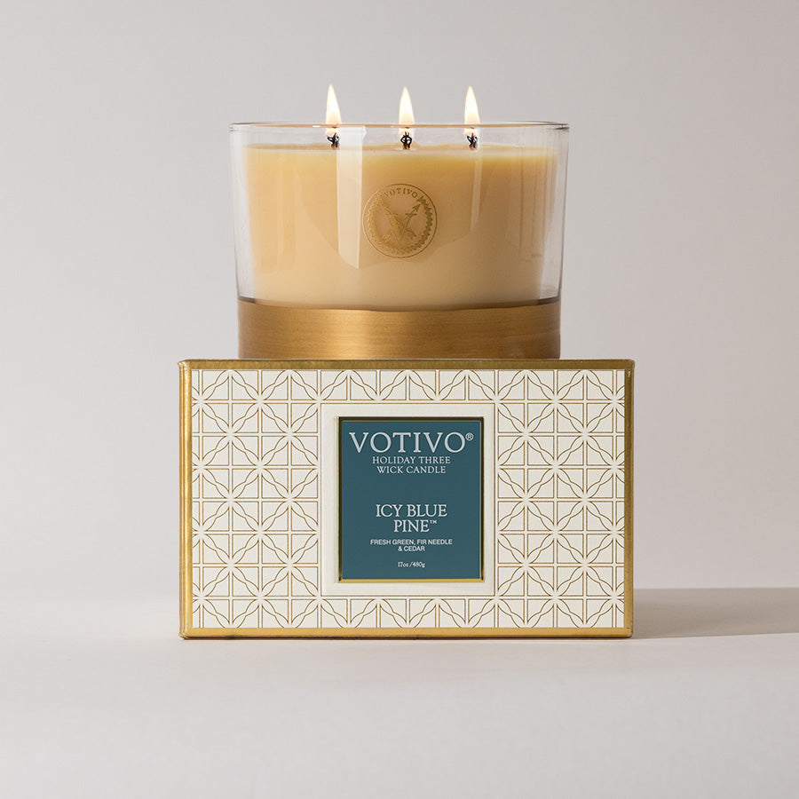 A beige Votivo Icy Blue Pine Holiday 3 Wick Candle sits atop its gold and white box. The label notes fresh citrus & Hermitage, 450g soy blend wax, with visible flames glowing from the top.