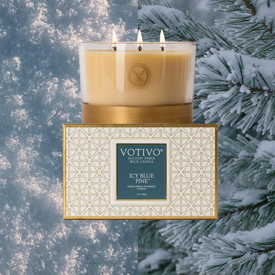 The Votivo Icy Blue Pine Holiday 3 Wick Candle, made from premium soy blend wax, sits lit atop its decorative box, framed by a snowy pine branch and a frosty blue backdrop.