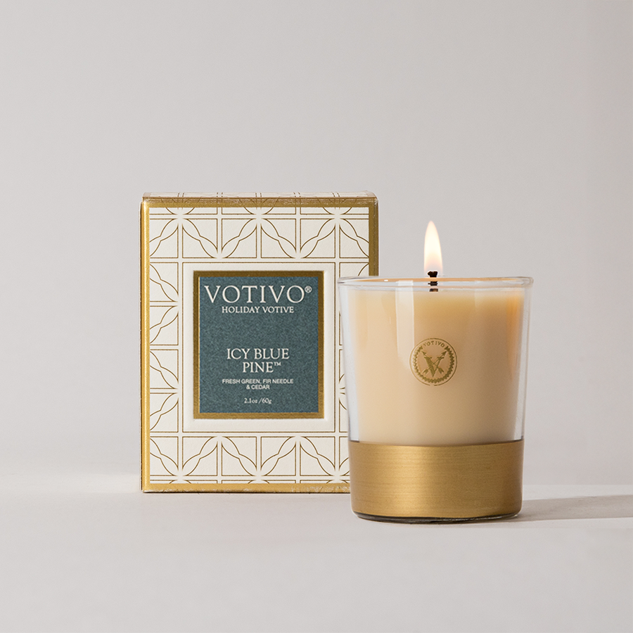 A lit Votivo Icy Blue Pine Holiday Votive made with soy blend wax sits in a glass holder with a gold base, displayed in front of its geometric gold and teal-accented box.