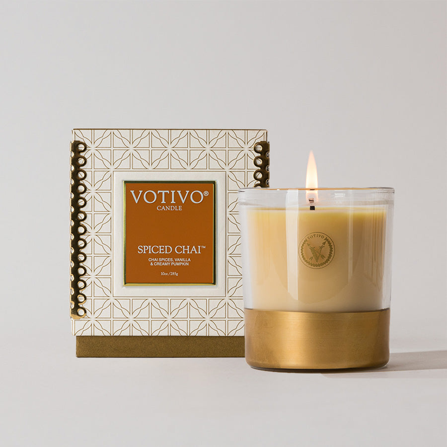 A lit Votivo Spiced Chai 10 oz. Holiday Candle with soy blend wax glows in a glass jar with a gold base, beside its decorative white and gold geometric box featuring an orange label.