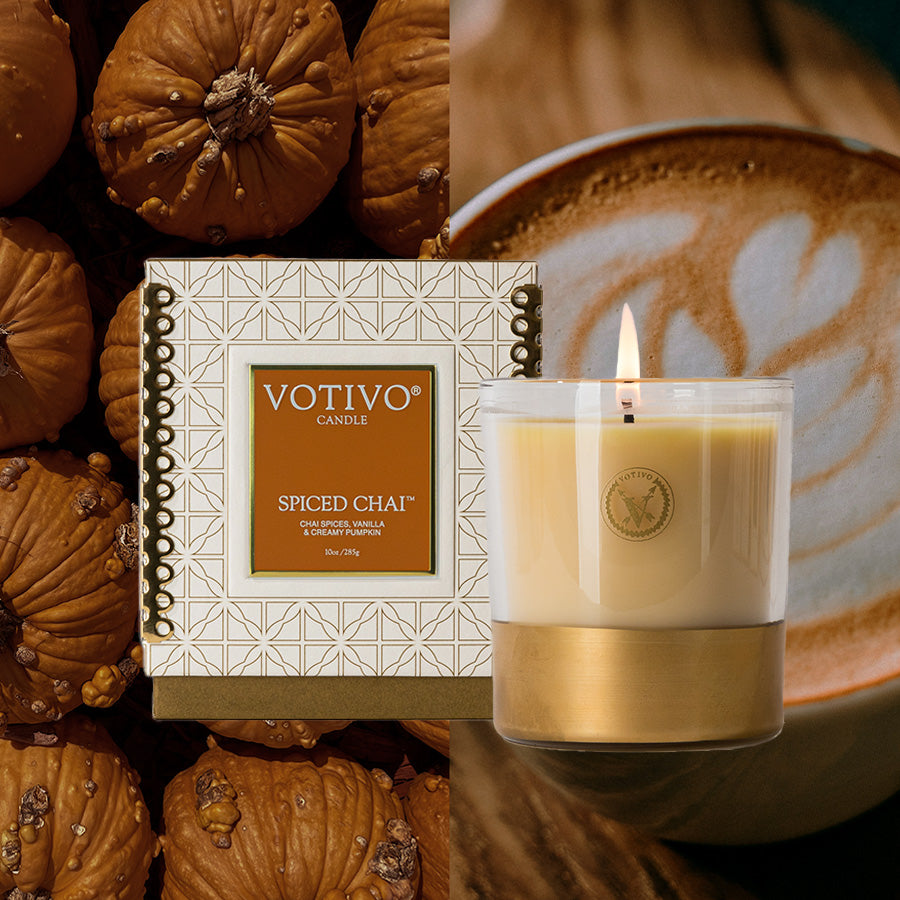 A Votivo Spiced Chai 10 oz. Holiday Candle sits among pumpkins, beside a burning soy blend wax candle in a glass holder and a foamy chai latte.
