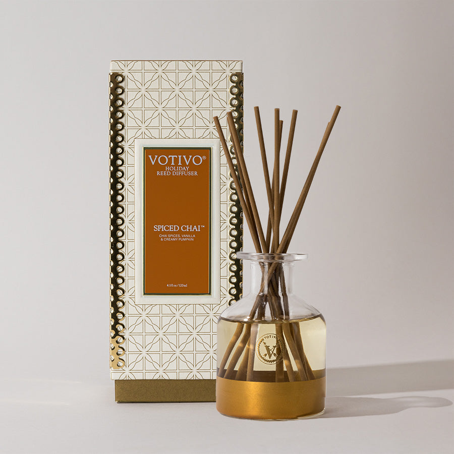 A Votivo Spiced Chai Holiday Reed Diffuser, with golden liquid and brown reeds in a glass bottle, stands in front of its decorative box displaying the product and scent name.