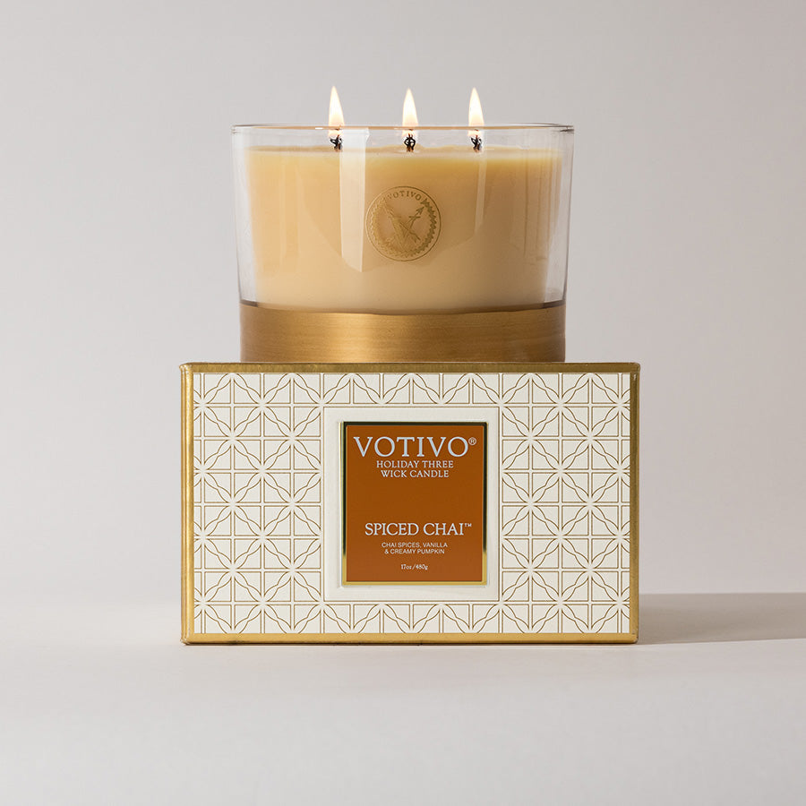 A Votivo Spiced Chai Holiday 3 Wick Candle, made with soy blend wax and featuring a tan color and white-and-gold geometric box, is lit and placed on display.