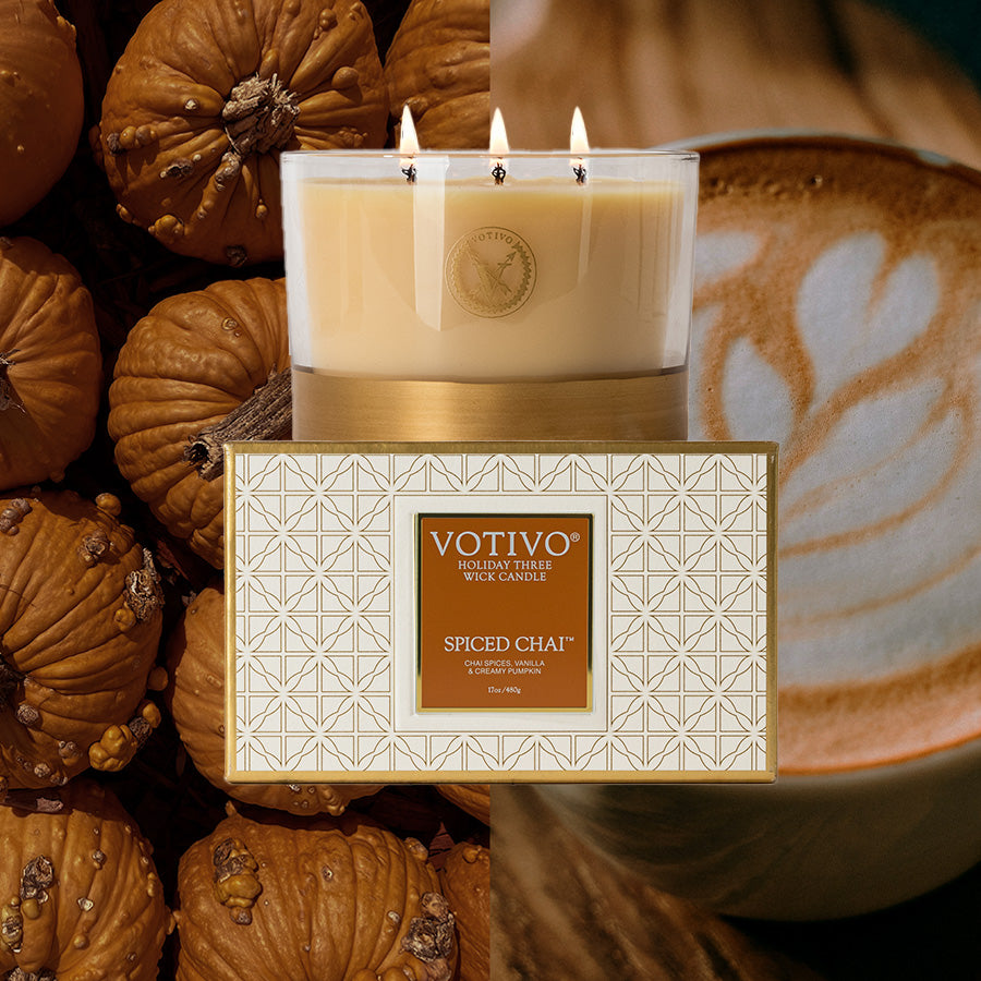 The Votivo Spiced Chai Holiday 3 Wick Candle sits atop its box, surrounded by small pumpkins and a frothy chai latte. Made with a soy blend wax, it fills your space with the cozy scents of the season.