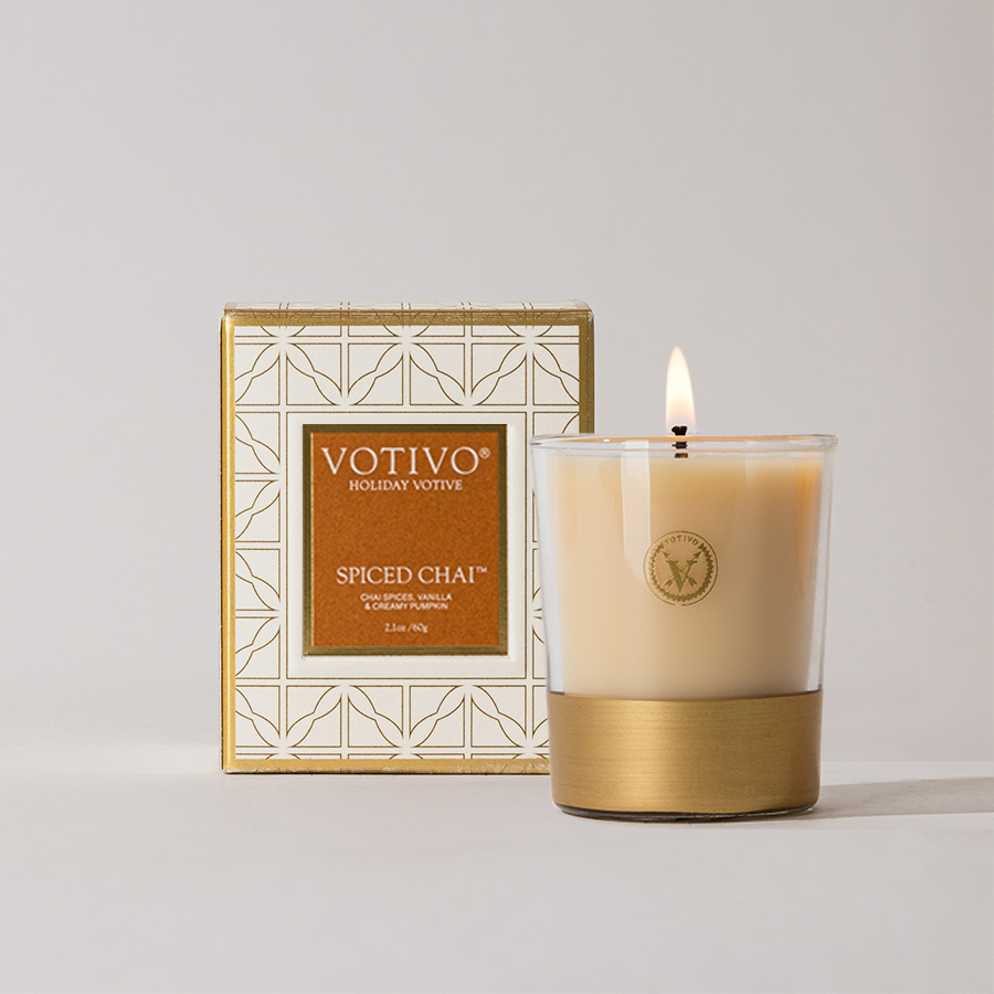 The Votivo Spiced Chai Holiday Votive, made from a soy blend wax in a glass container with a gold base, is shown lit in front of its decorative box featuring geometric patterns and gold accents.