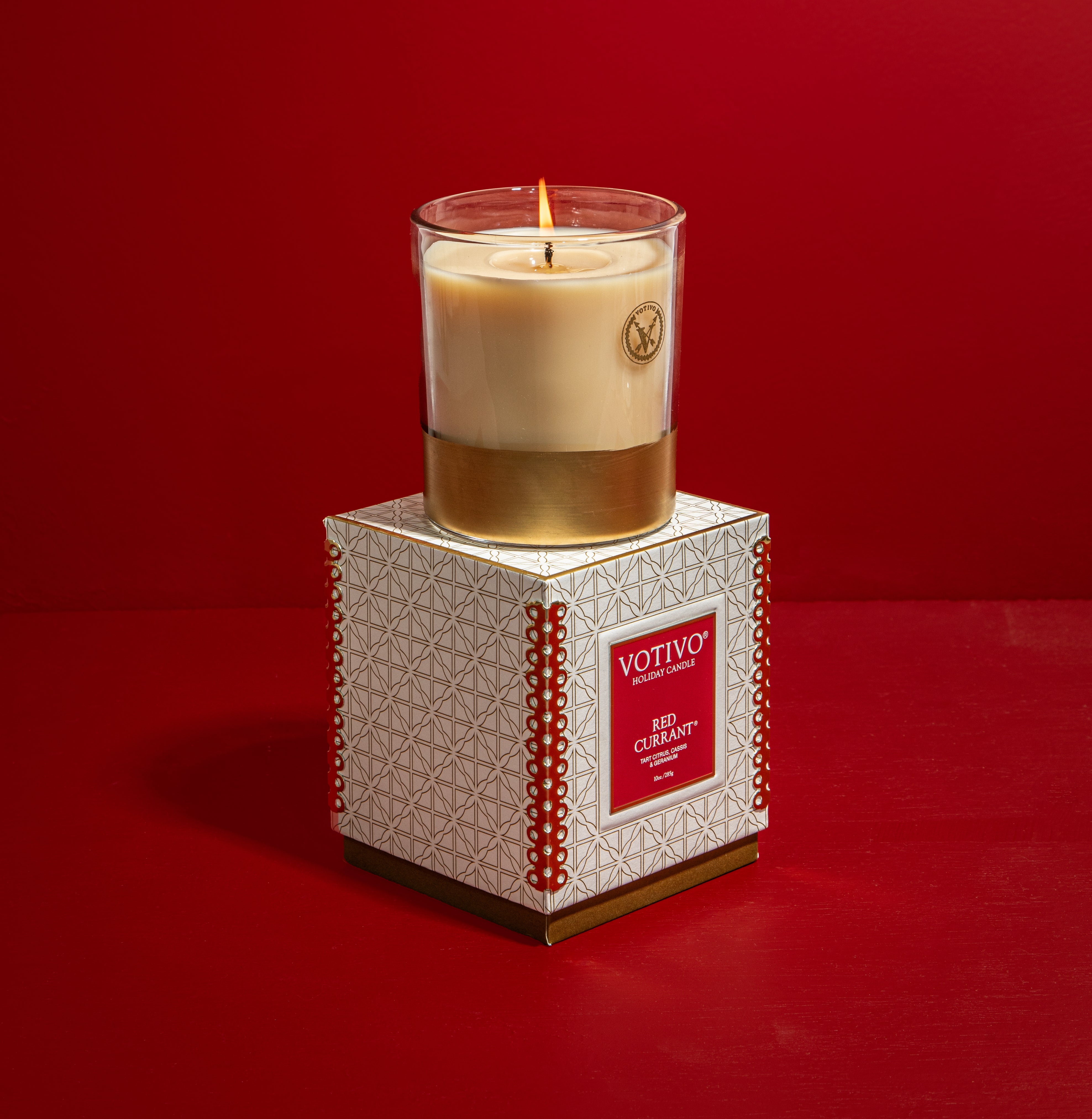 A Votivo Red Currant 10 oz. Holiday Candle with soy blend wax glows in a glass jar, displayed on its decorative white and gold box against a solid red background.