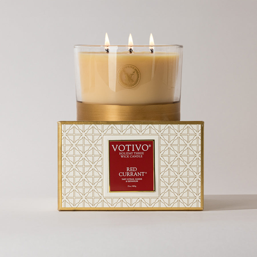 A lit Votivo Red Currant Holiday 3 Wick Candle rests on a decorative box with a gold geometric design and red label, filling the air with its vibrant red currant scent from this premium soy blend candle.