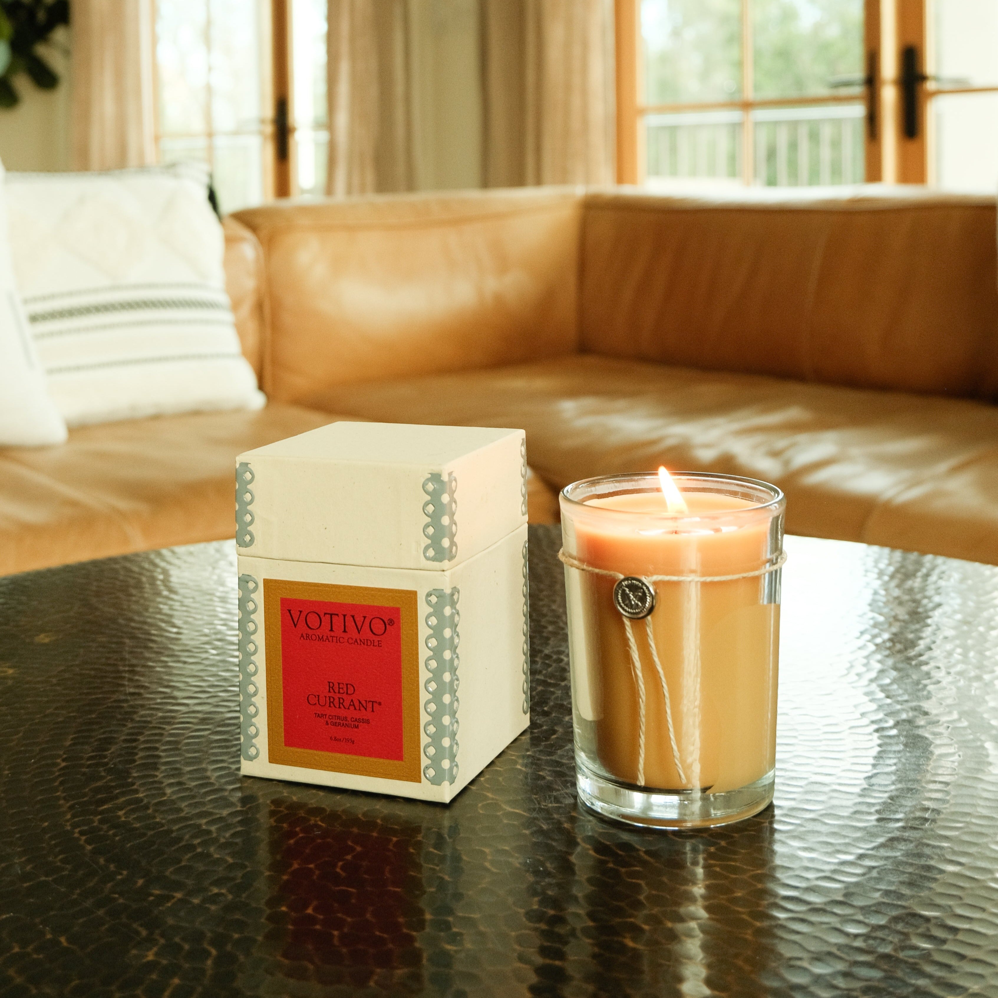 A Votivo Red Currant 6.8 oz. Aromatic Candle glows beside its box on a round, dark table in a cozy living room featuring a tan leather sofa and a window in the background.