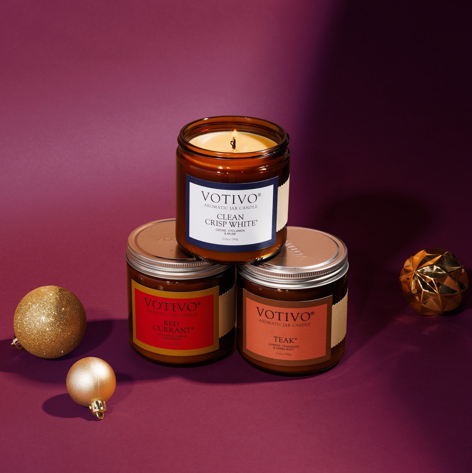 Three Votivo Limited Edition Candle Set – Red Currant, Clean Crisp White & Teak Large Amber Jars are arranged on a maroon surface with holiday ornaments, one candle lit and stacked atop the others.