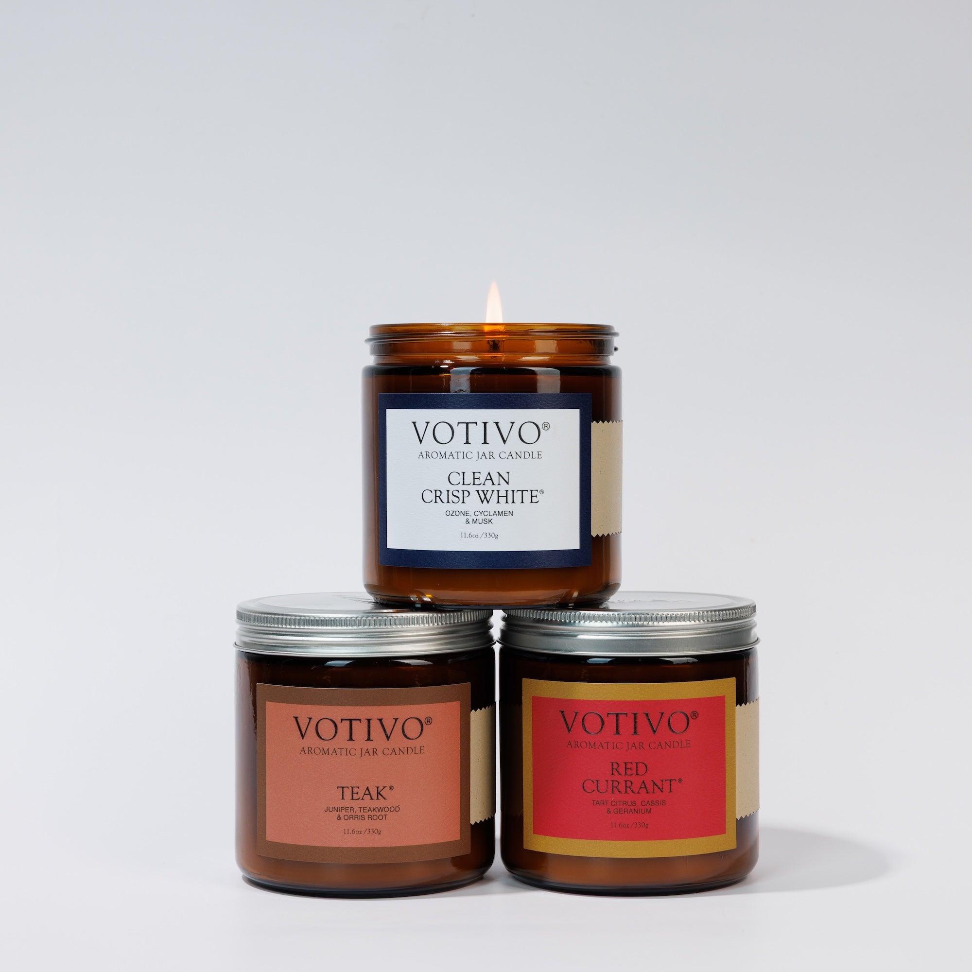 Votivo’s Limited Edition Candle Set features three large amber jars—Red Currant, Clean Crisp White (lit on top), and Teak—arranged in a pyramid for a striking display.