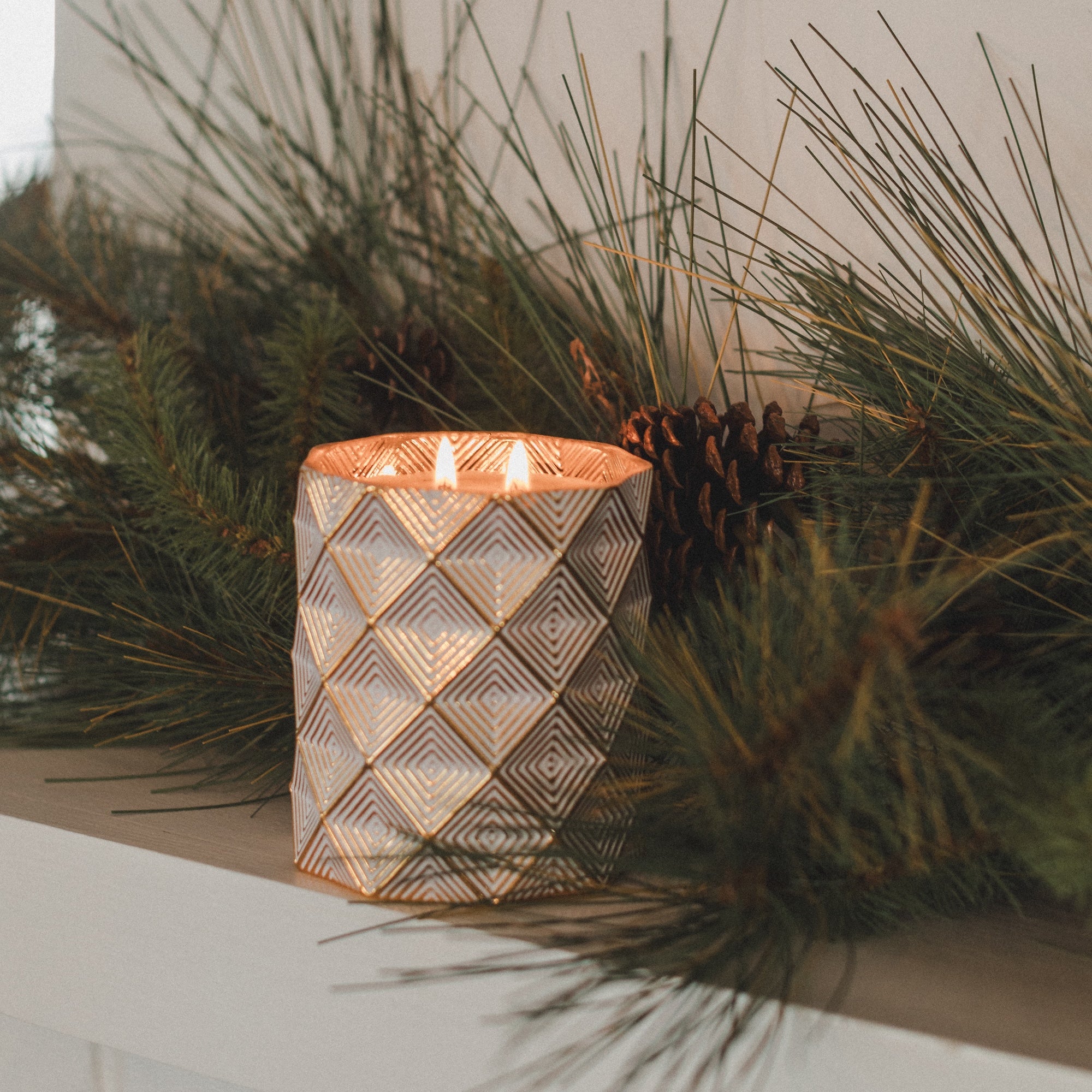 The Votivo Icy Blue Pine Holiday Decorative Candle, set in a patterned holder and surrounded by pine branches and pinecones on a white surface, creates a cozy, festive vibe.