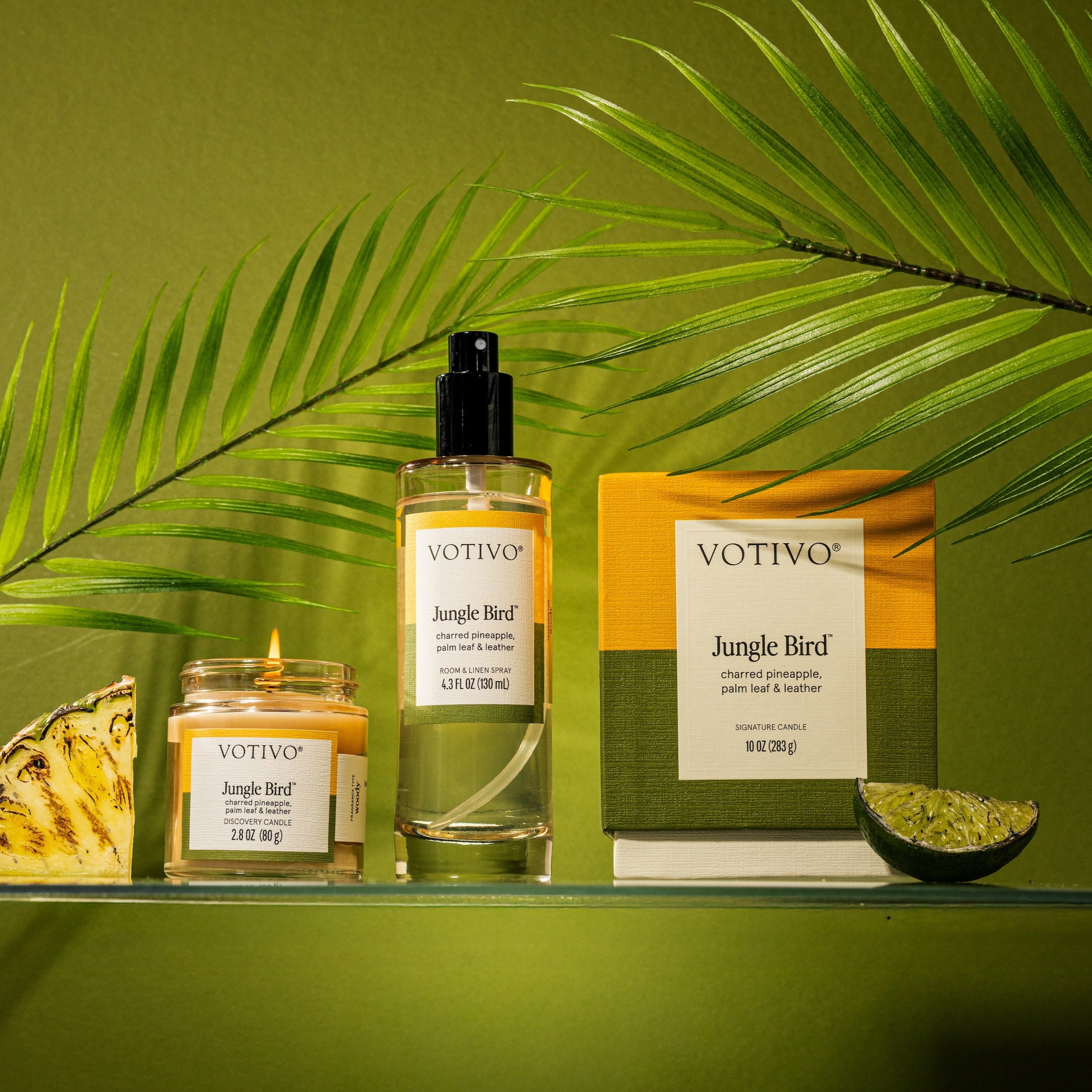 Votivo’s Jungle Bird Discovery Candle, room spray, and box are displayed with tropical leaves, pineapple, and lime on green, emphasizing the candle’s long-lasting fragrance and soy blend.