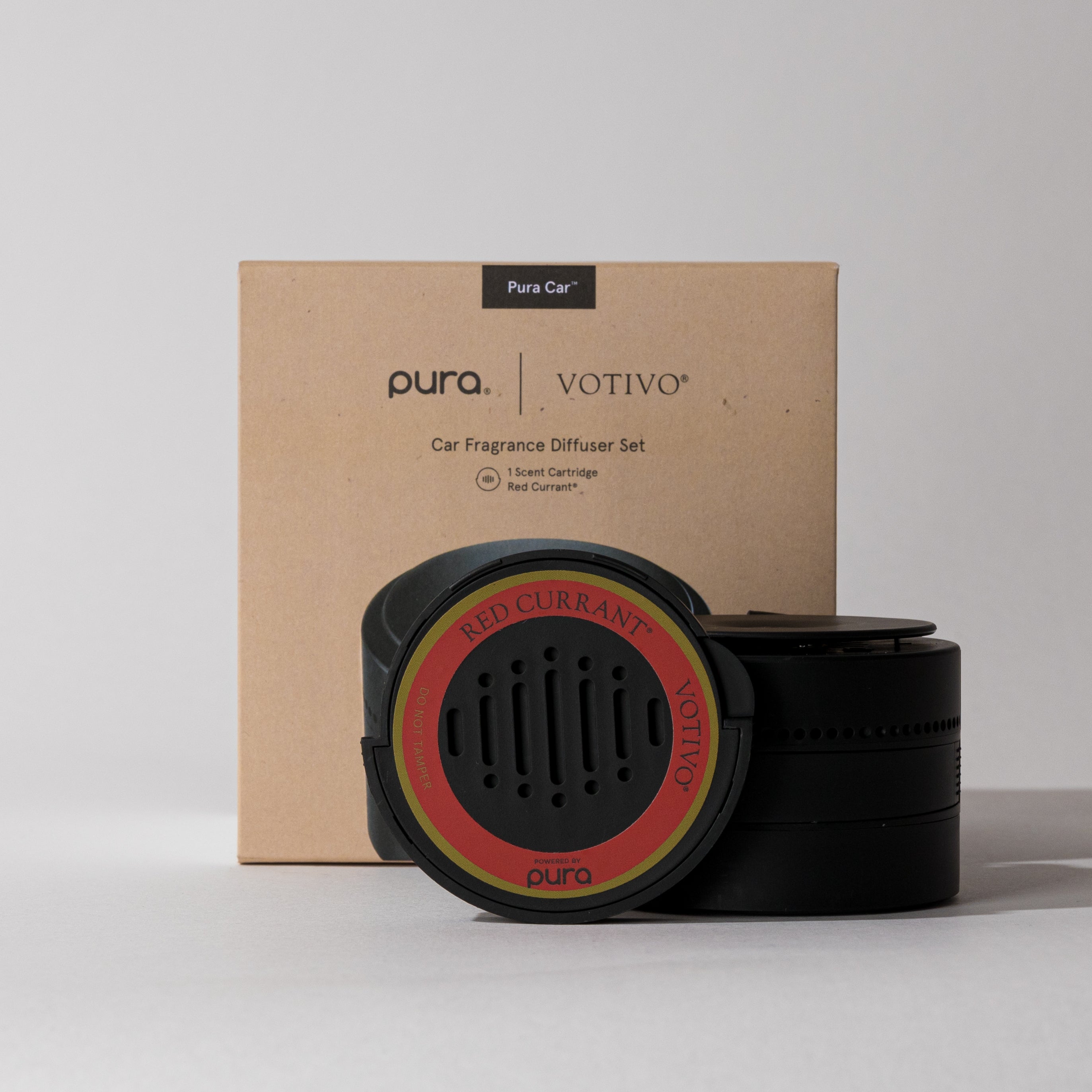 The Votivo Red Currant Pura + Votivo Car Diffuser Set is shown in front of its brown box. The black diffuser, with a red label, offers a hands-free car scent experience.