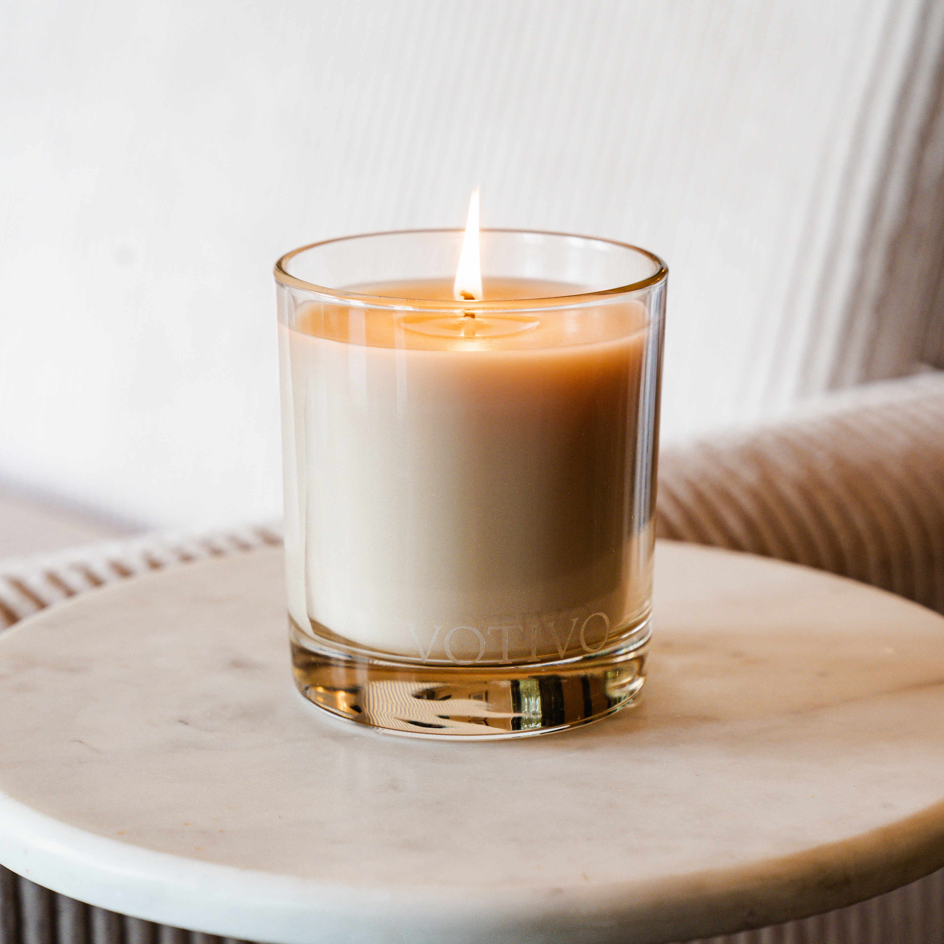 A lit Votivo Red Currant 10 oz. Signature Candle made from soy blend wax in a clear glass holder sits on a round white marble table, set against a softly blurred beige fabric background.