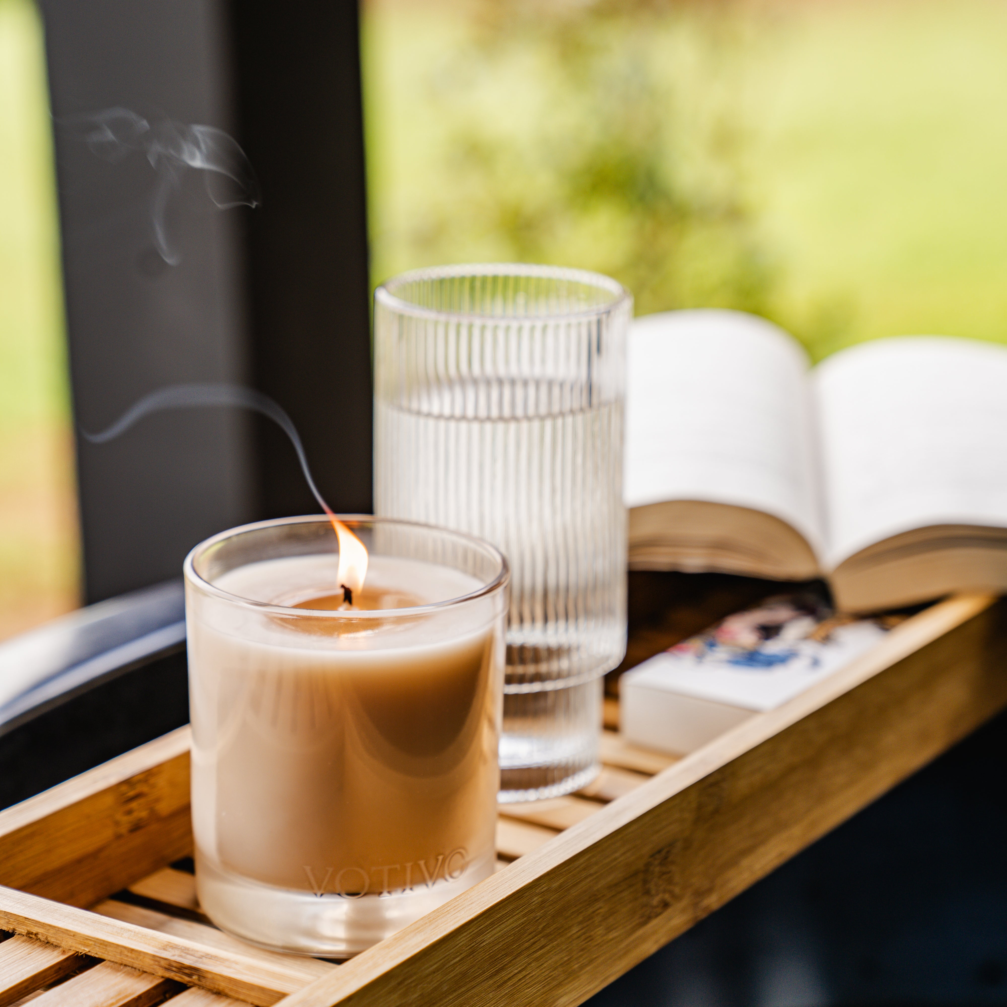 A Votivo Red Currant 10 oz. Signature Candle made from soy blend wax sits on a wooden tray alongside a glass of water, a book, and matches, while green foliage blurs in the background and red currant aroma fills the air.