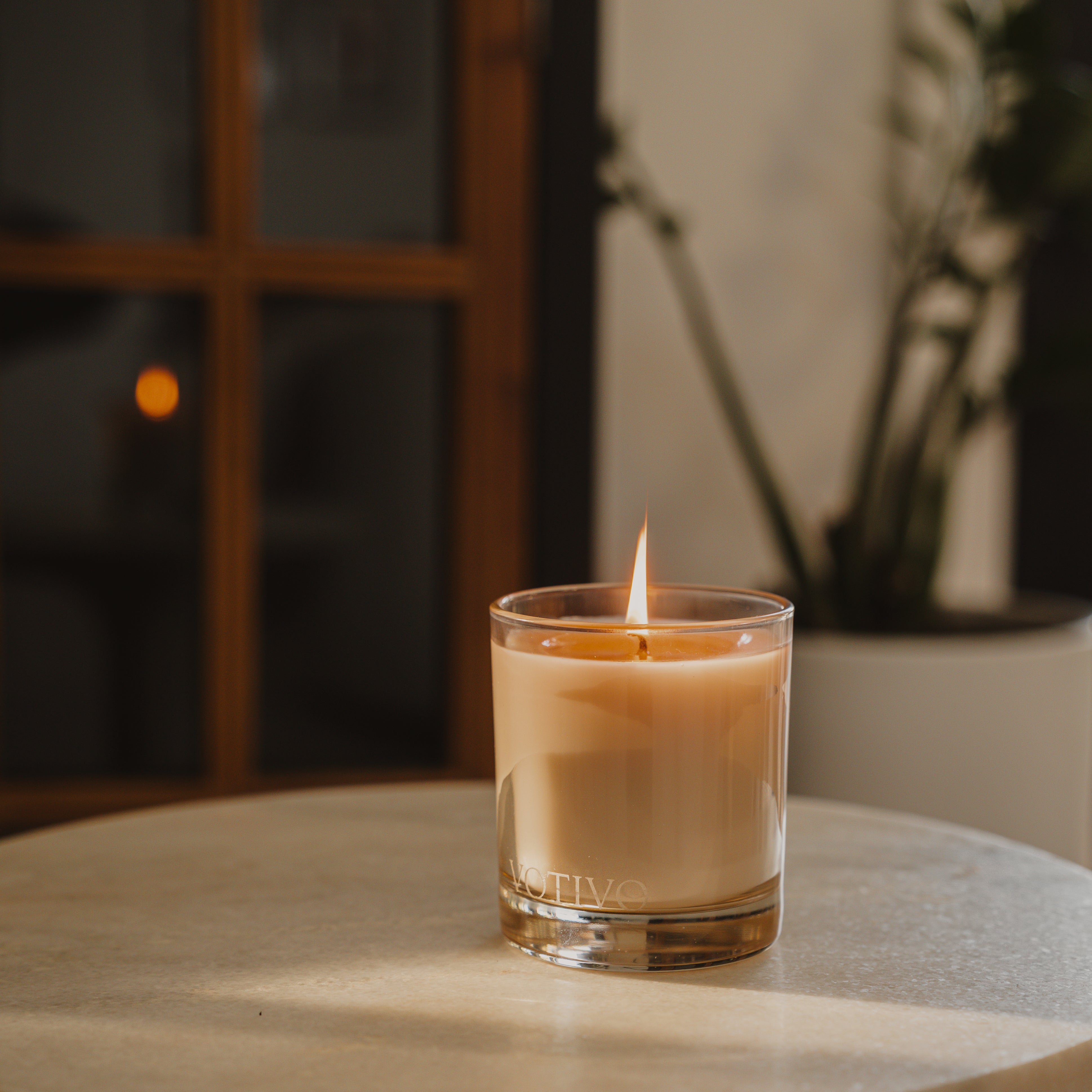 A lit Votivo Island Grapefruit 10 oz. Signature Candle made from soy blend wax sits on a light round table, with a blurred plant and wooden window frame in the background. Warm, soft lighting creates a cozy feel.